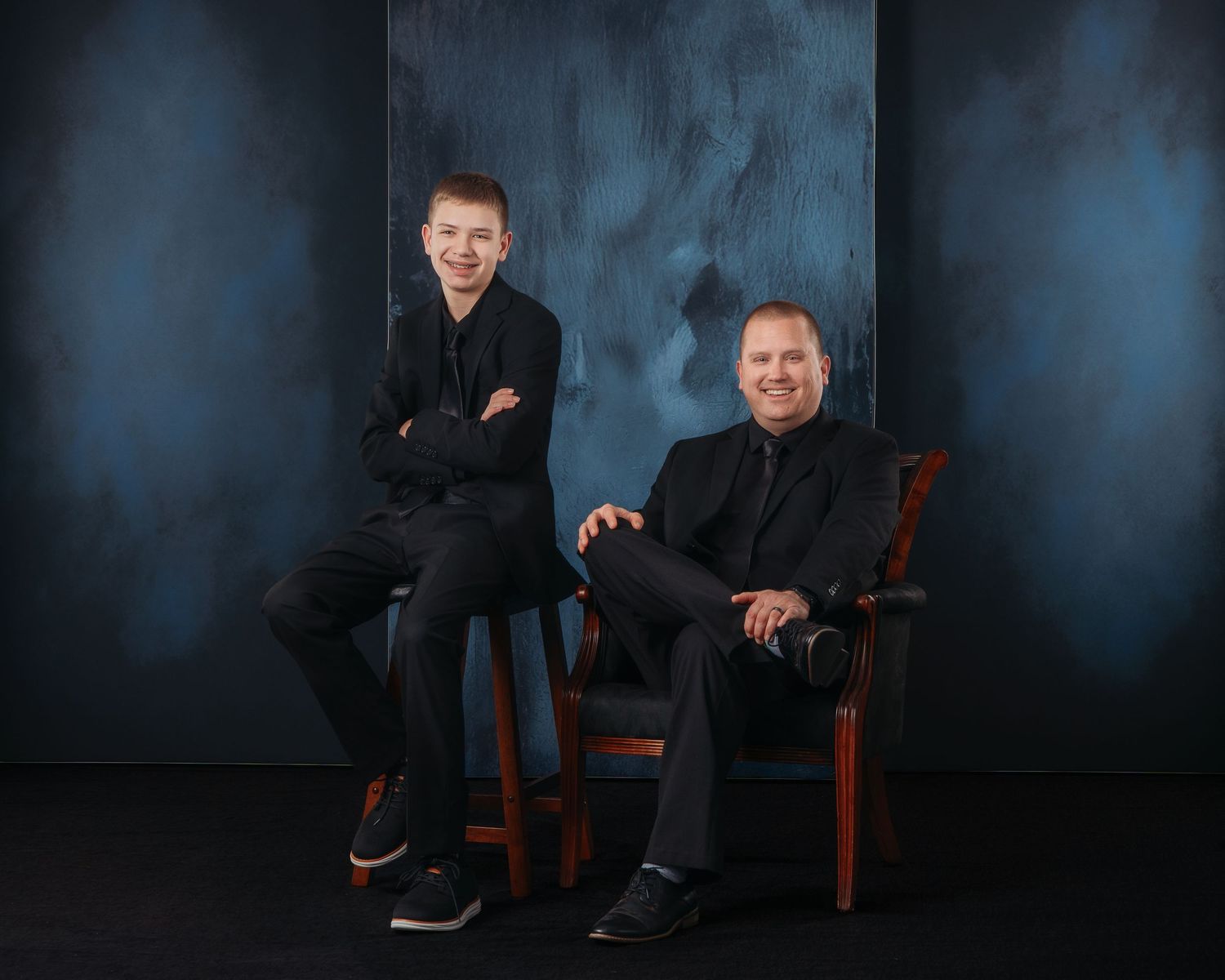 Two men in black attire pose for a professional portrait against a dark blue studio background.