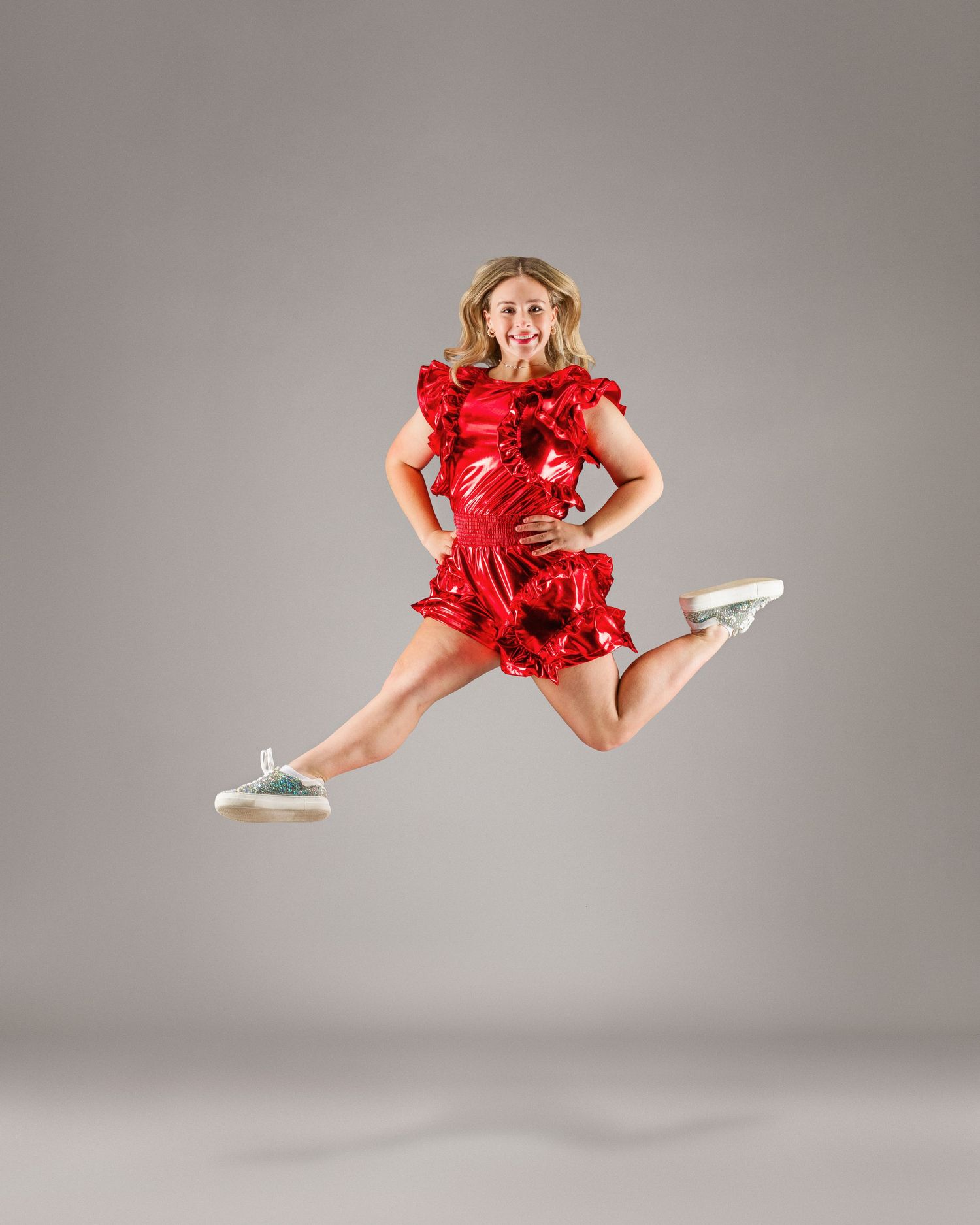 Young woman in shiny red ruffled dress leaping mid-air with a smile in a professional studio photo.