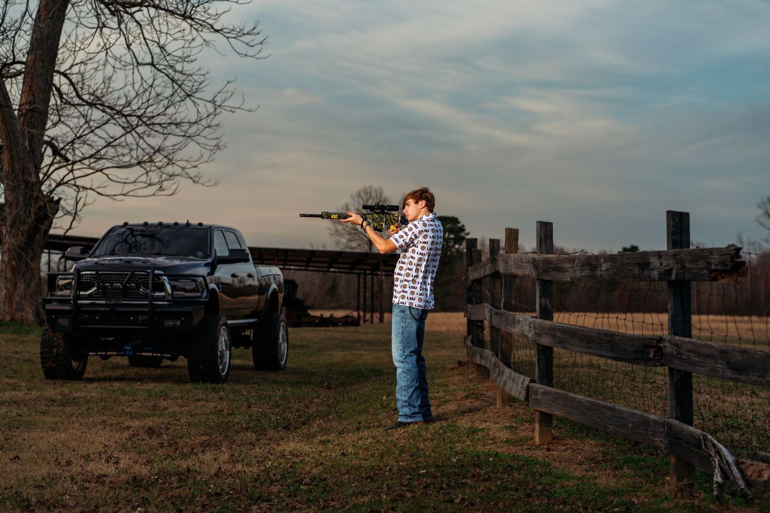 Man aiming a rifle near a black truck by a wooden fence on a farm at dusk.