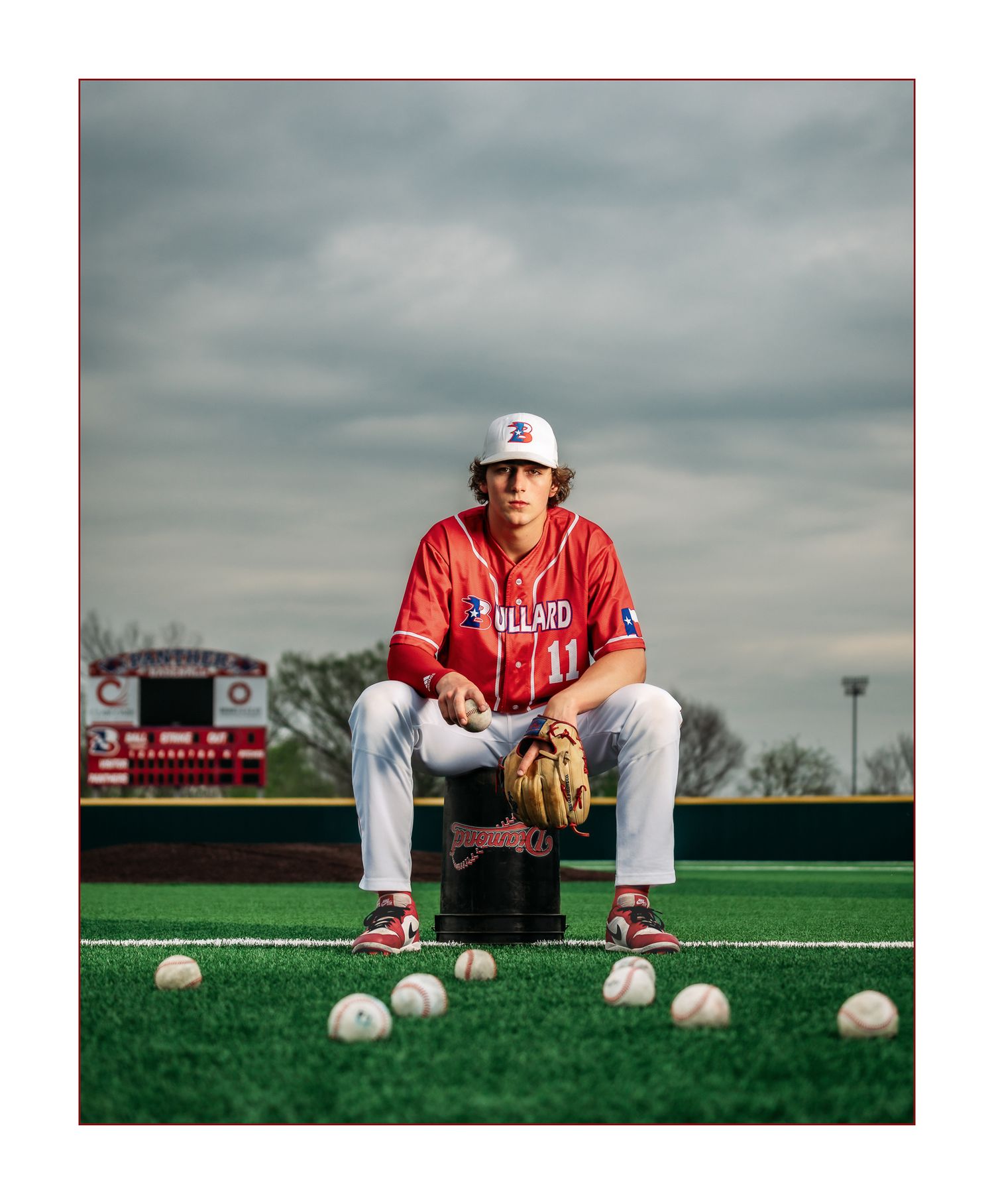 Baseball player in red Bullato uniform sitting on a bucket surrounded by baseballs on a green field.