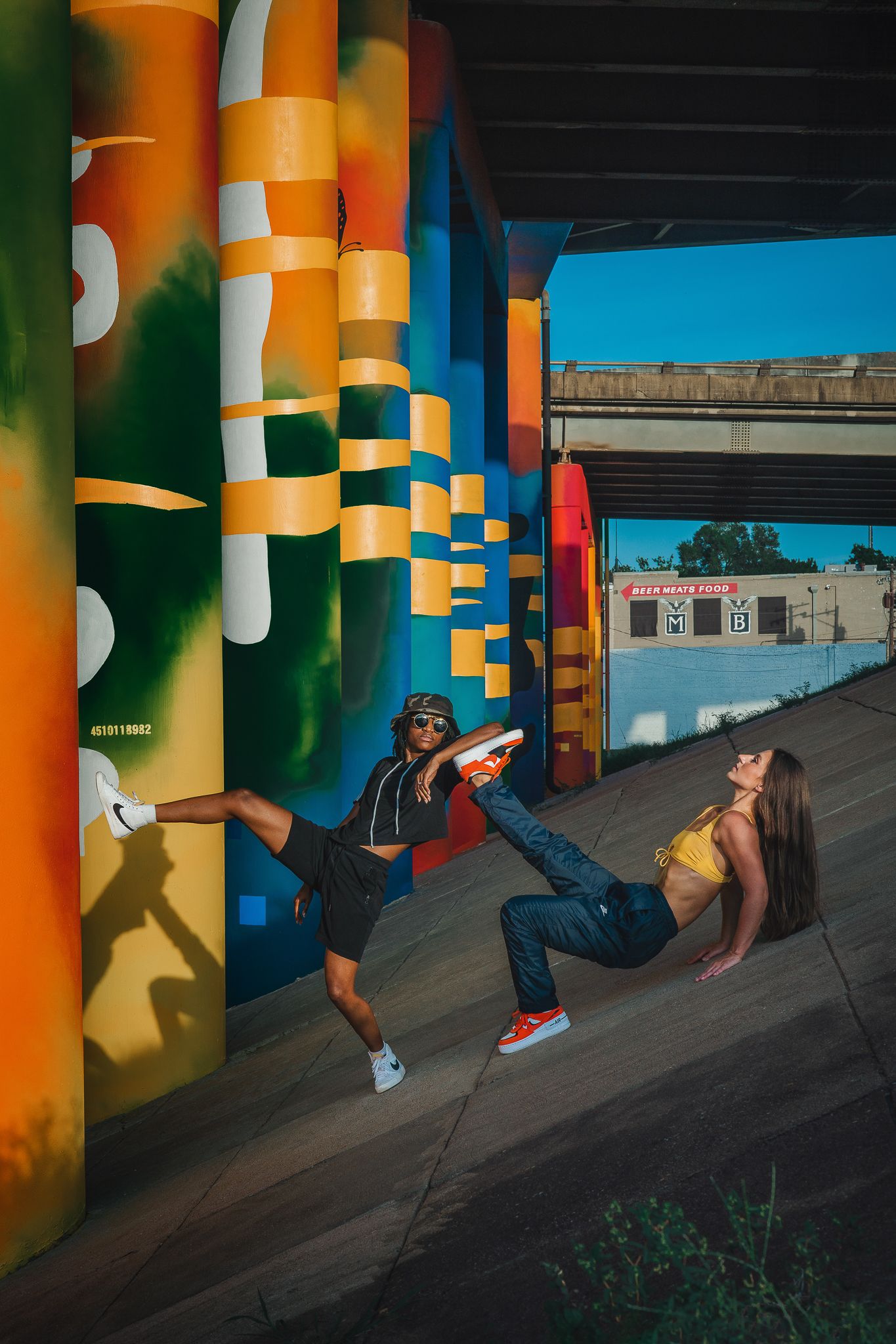 Two dancers posing dynamically beside colorful painted columns under a bridge.