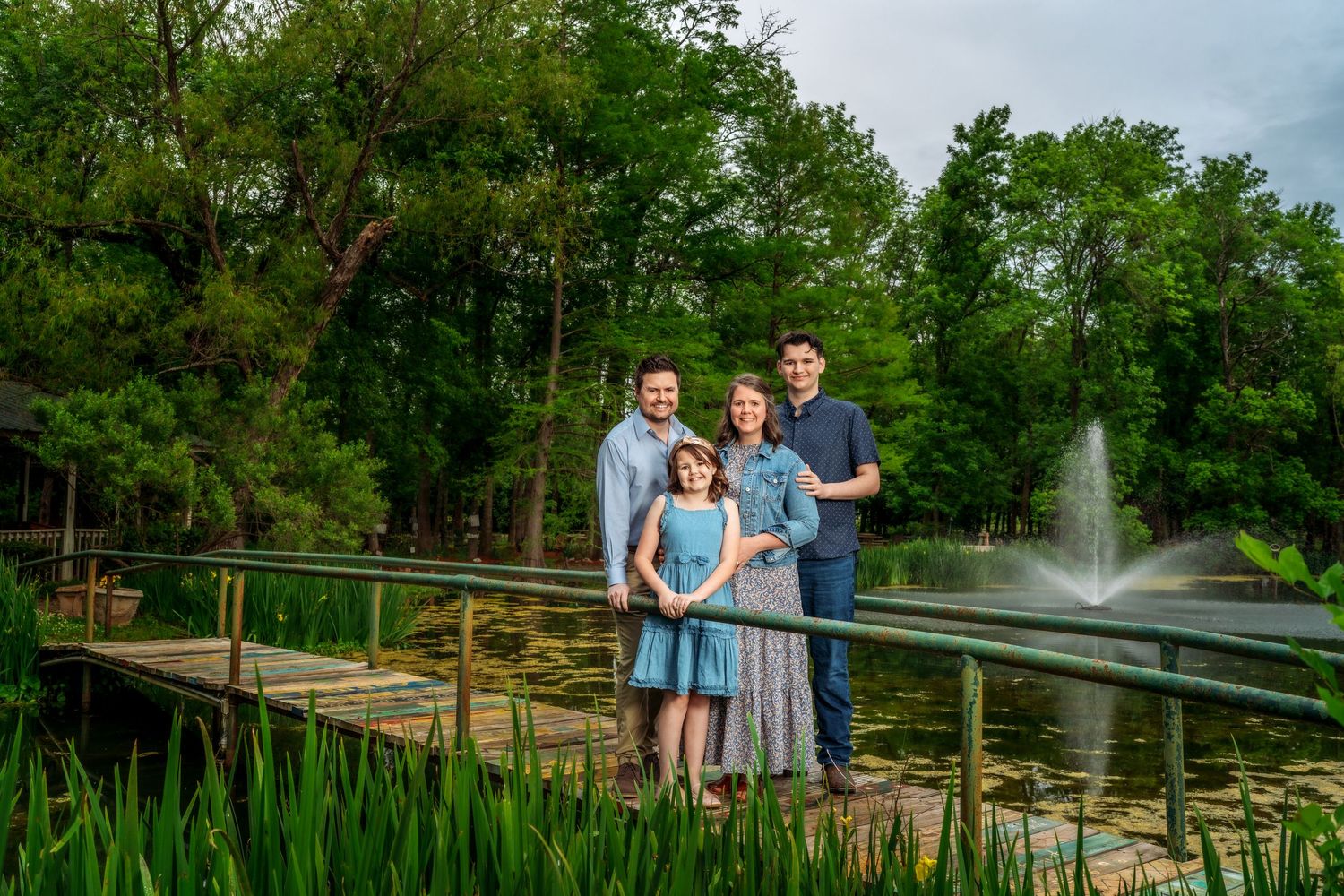 Family of four posing on a wooden dock surrounded by a pond with a fountain and lush green trees.