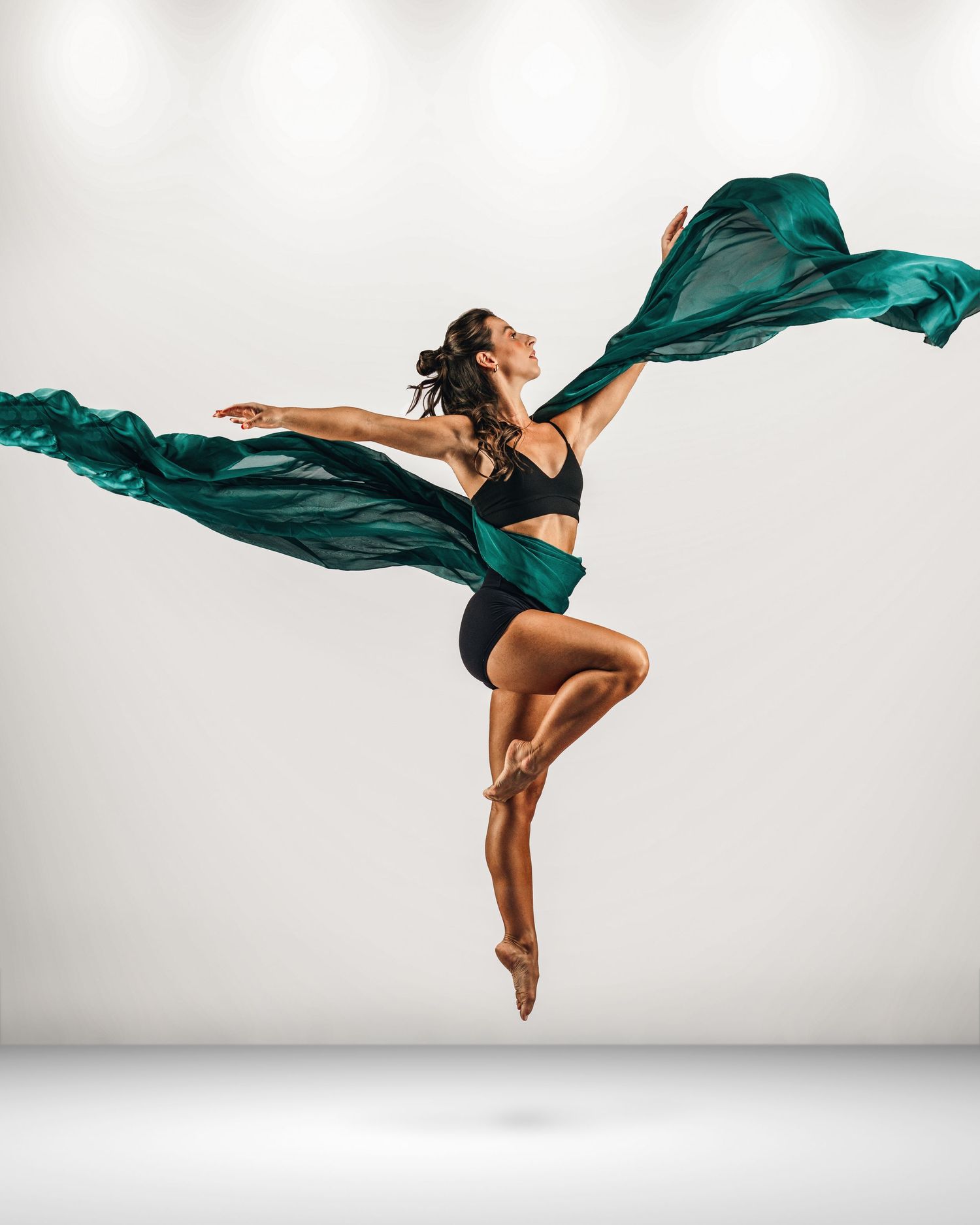 Dancer leaping mid-air in black outfit with flowing teal fabric against white background.