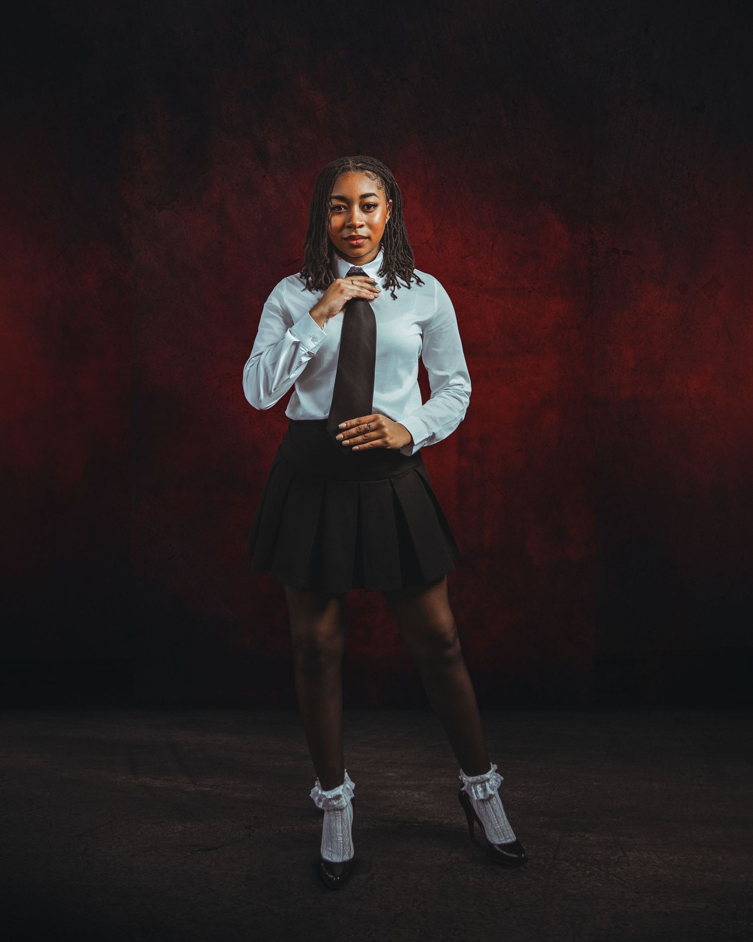 Young woman in white shirt, black pleated skirt, tie, and frilly socks poses confidently against dark red backdrop.