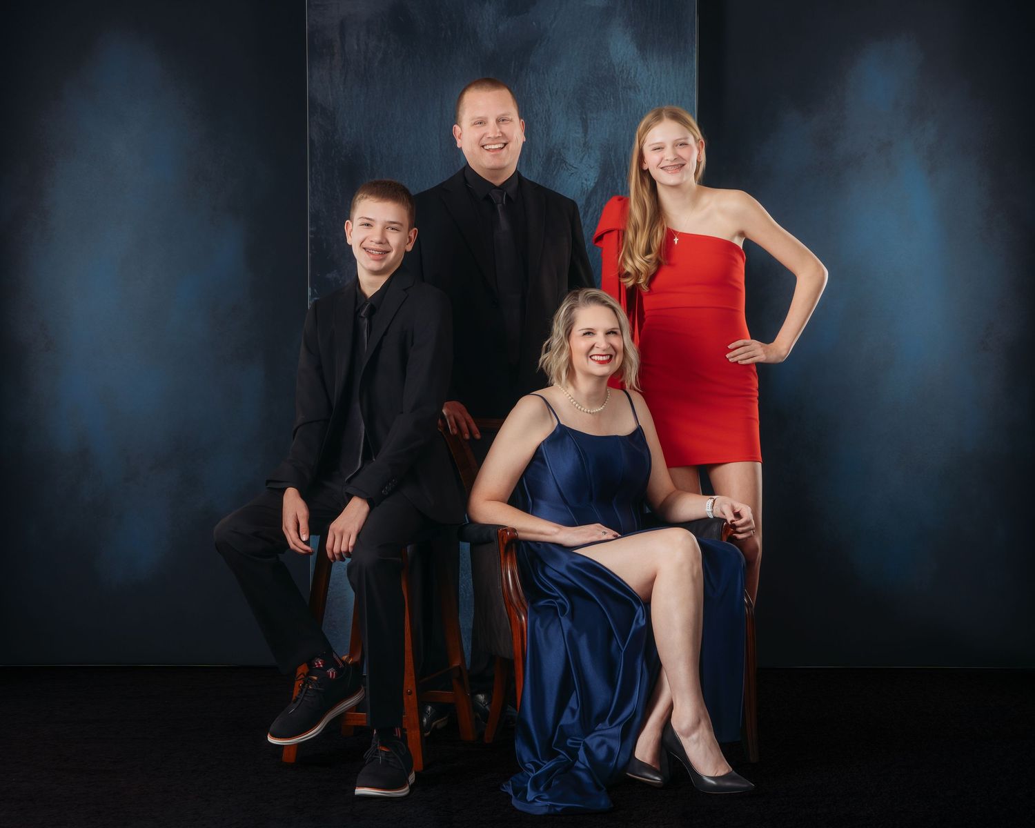Family of four posing for a formal portrait, wearing elegant attire in red, blue, and black, against a dark blue backdrop.