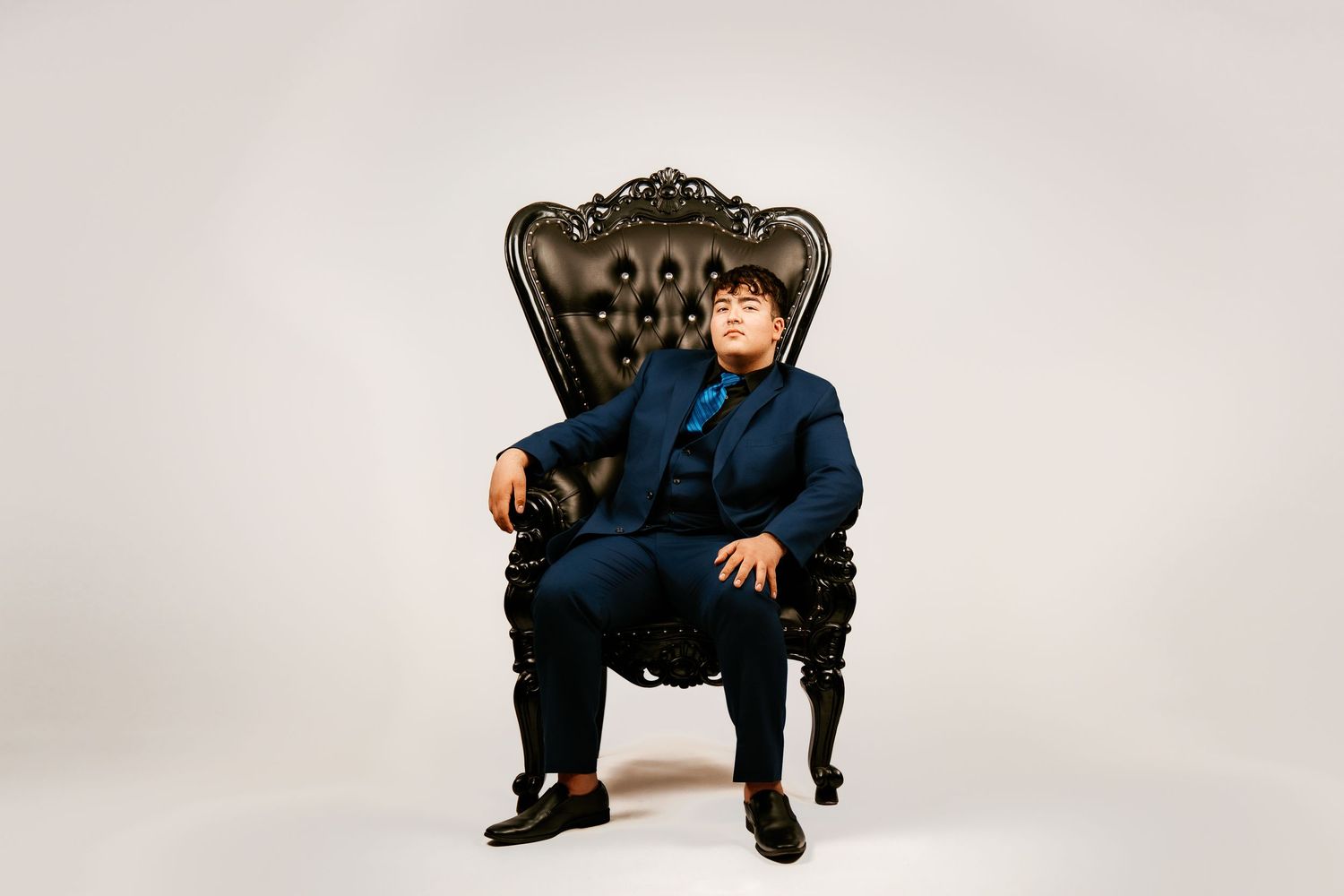 Young man in navy blue suit seated on ornate black throne chair against white background.