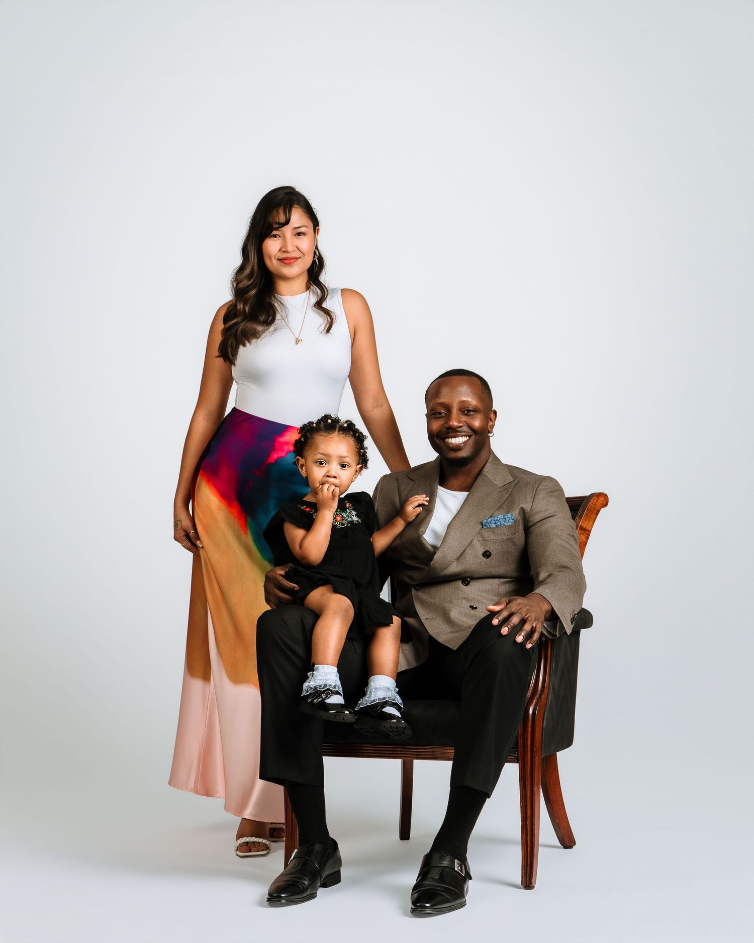 A joyful family portrait featuring a woman, man in a suit, and toddler posing together in a studio setting.