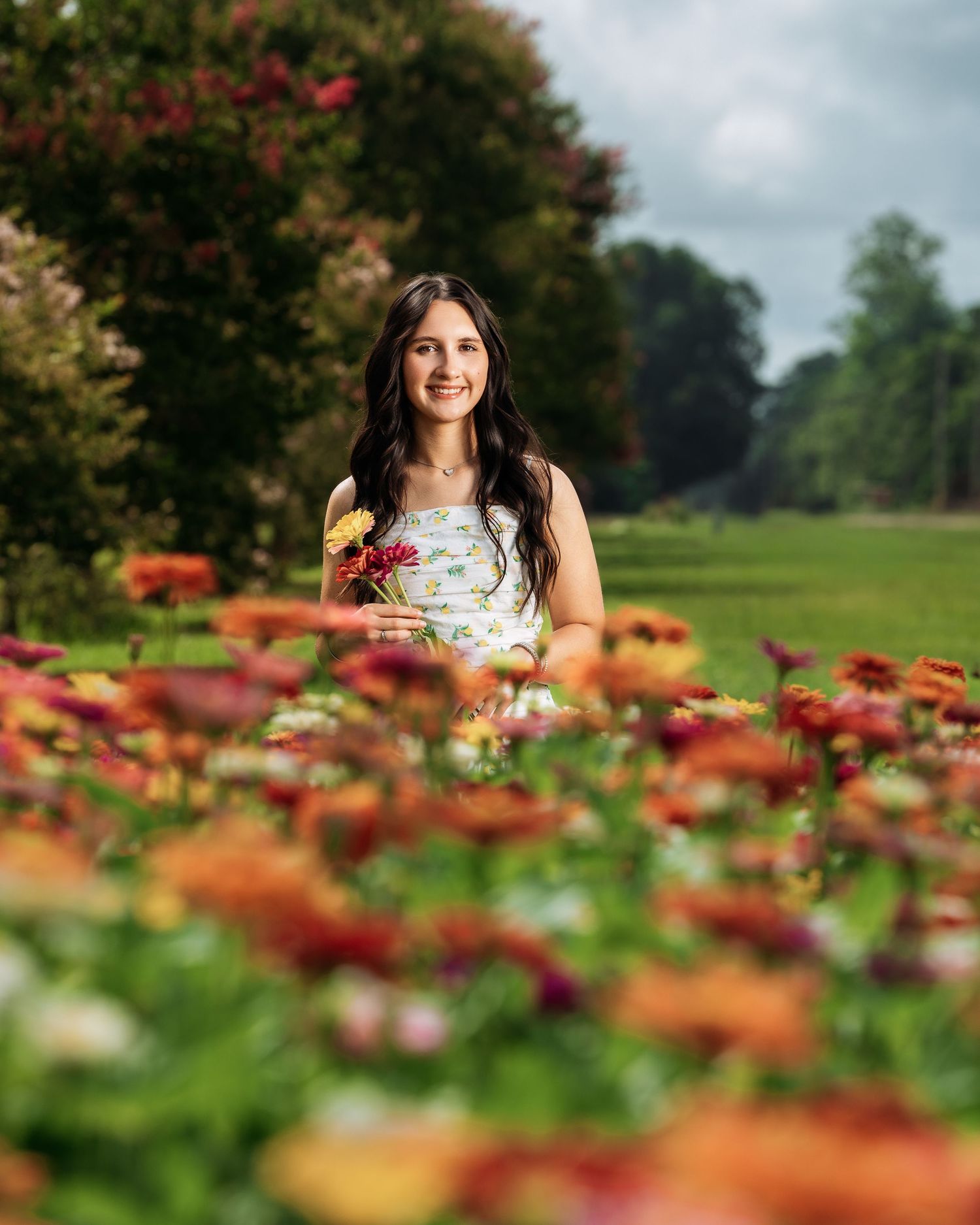 A young woman smiles while sitting among vibrant orange and red zinnia flowers in a garden.