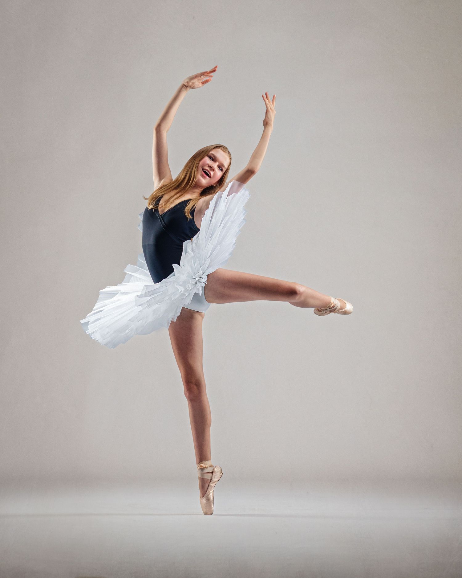 Smiling ballerina in white tutu and black leotard performs an arabesque on pointe in a studio.