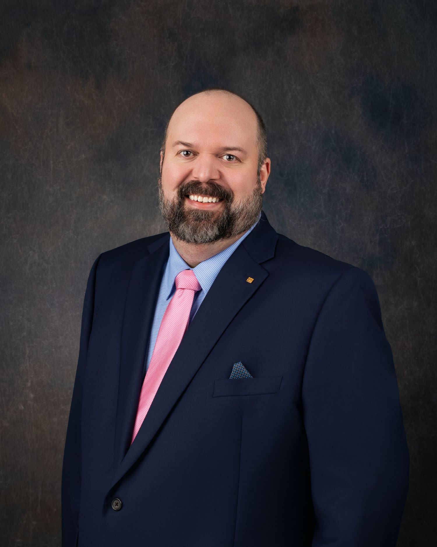Professional headshot of a smiling bald man with a beard wearing a navy suit and pink tie.