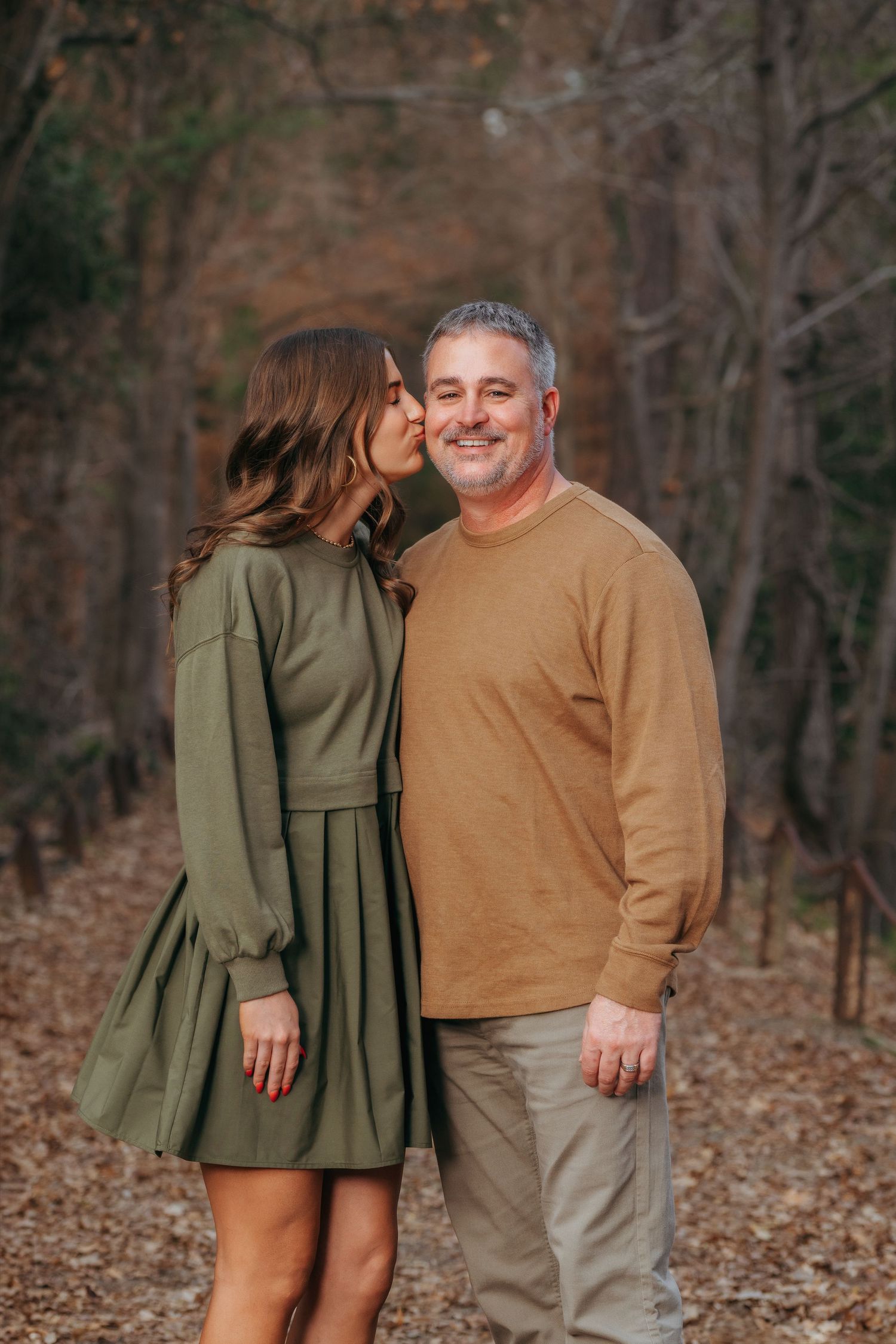Young woman in green dress giving a kiss on the cheek to smiling man in tan sweater on a forest path.