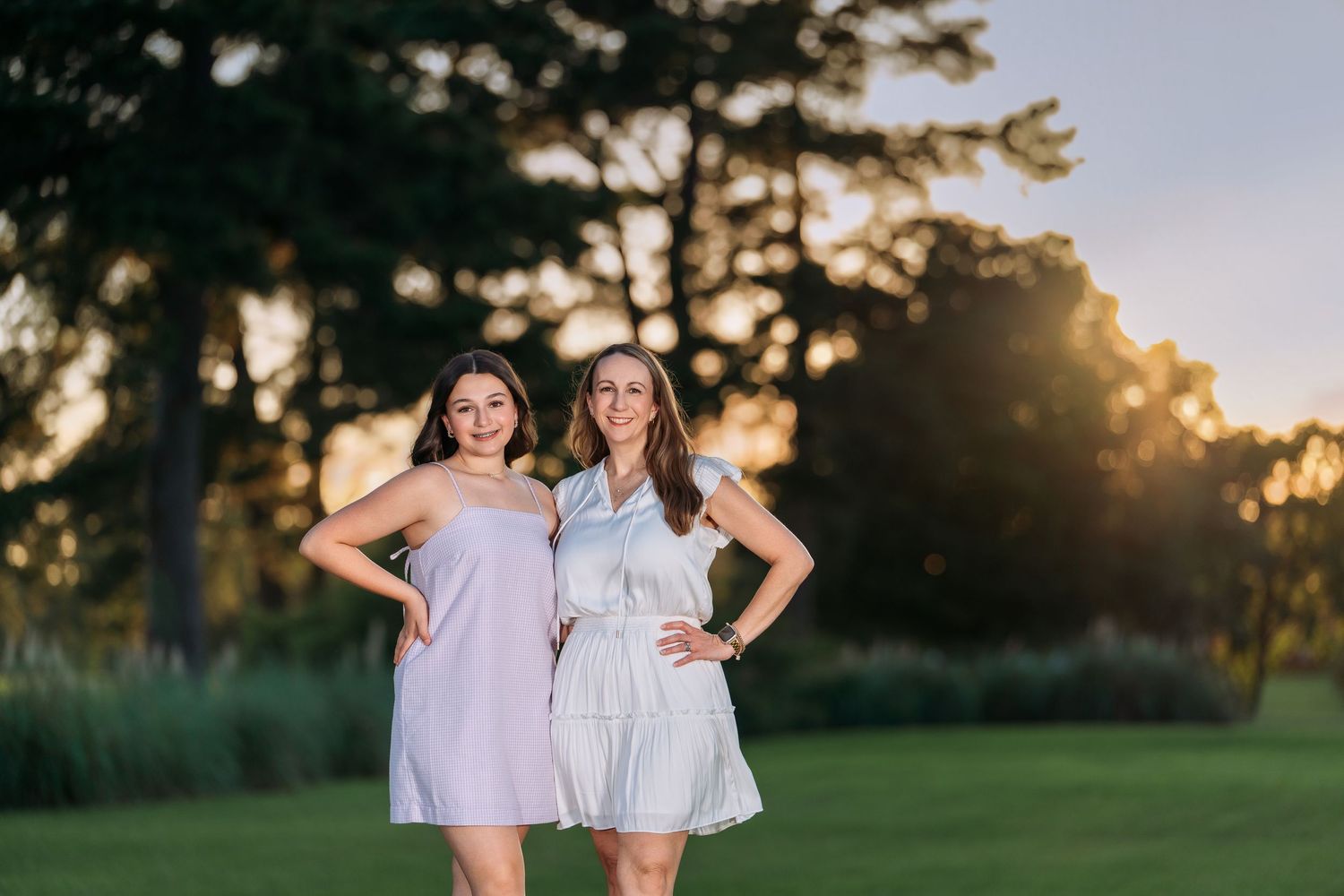 Two women in light-colored dresses pose with hands on hips on a sunlit green lawn.