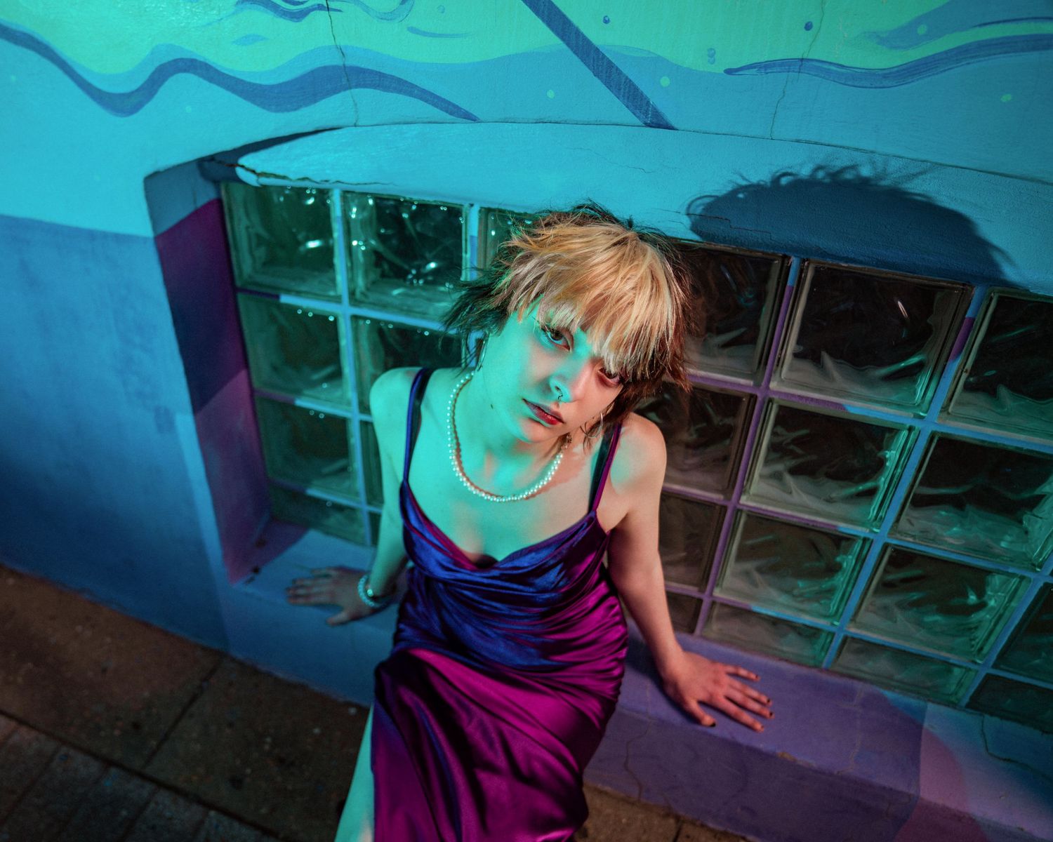 Blonde woman in purple satin dress poses against teal wall with glass block window under dramatic blue and pink lighting.