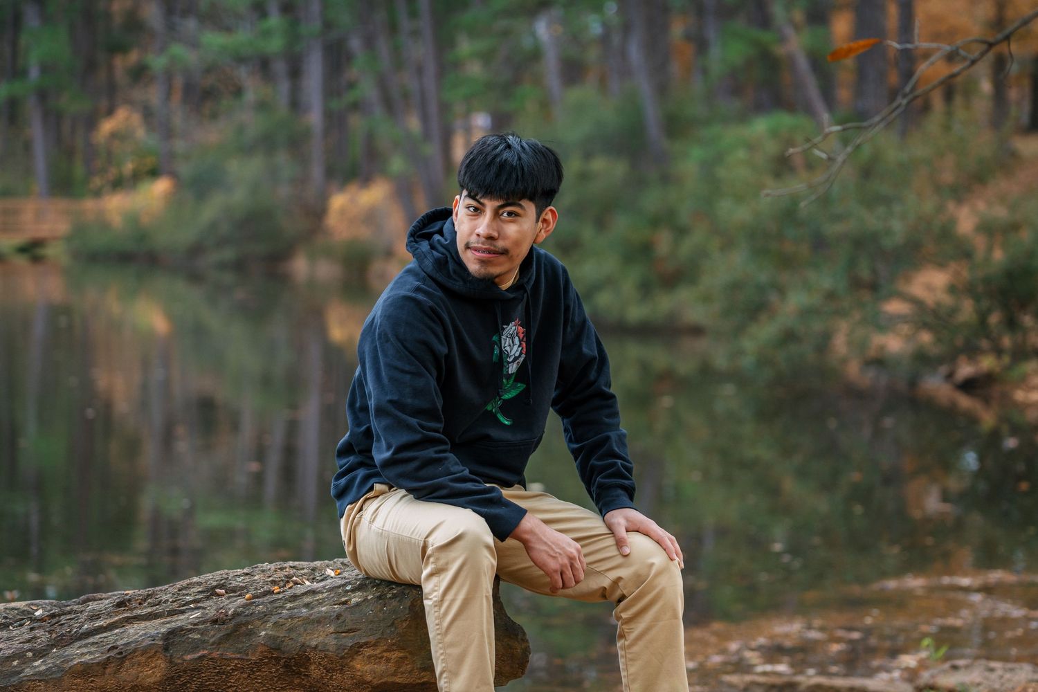 Young man sits on fallen log beside forest pond in casual outdoor senior portrait.