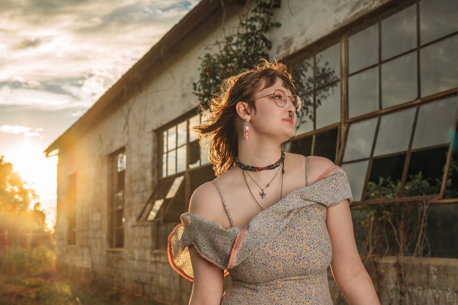 Young woman in floral off-shoulder dress posing outdoors at golden hour near rustic building.