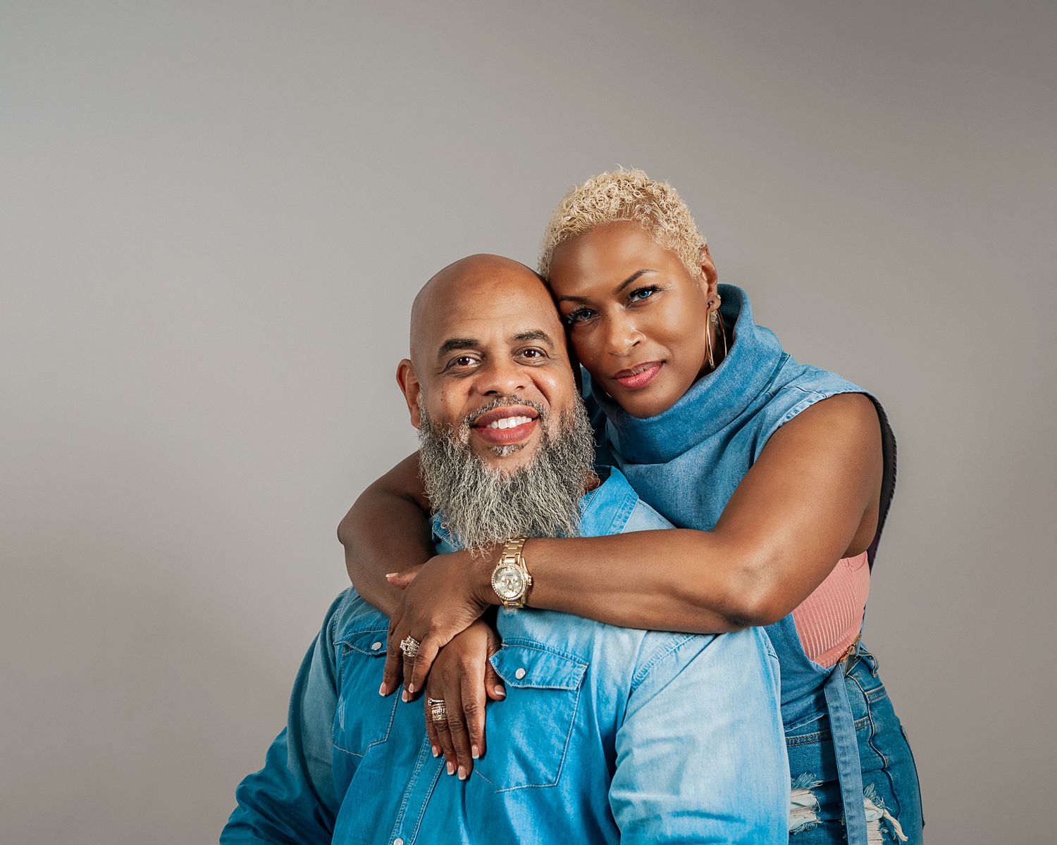 Smiling couple embracing warmly in matching denim outfits on gray background.