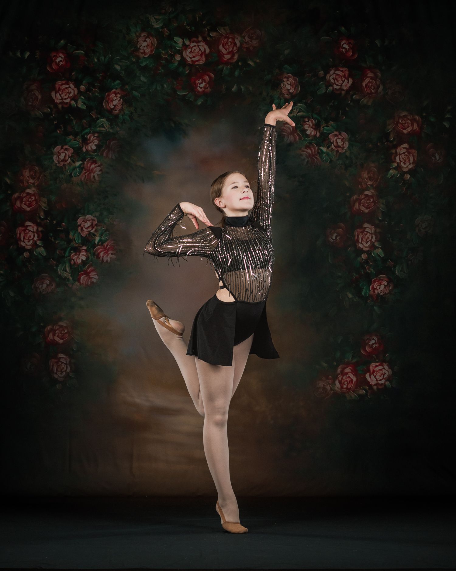 Young dancer in black sequin leotard performing arabesque before floral rose arch backdrop.