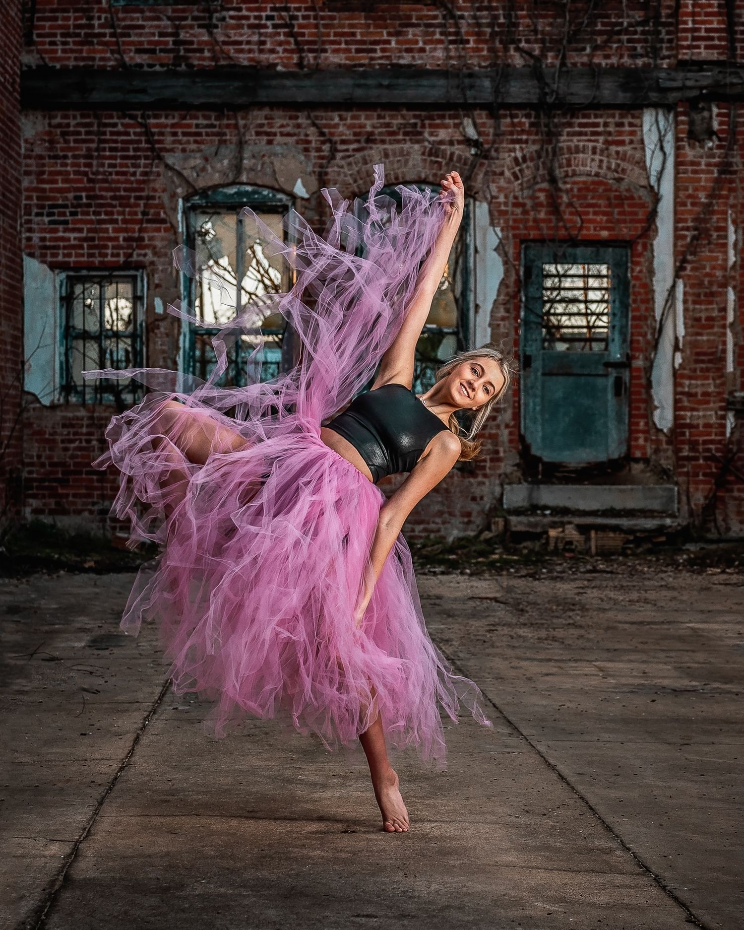 Ballerina in black leotard and flowing pink tulle skirt performs dramatic dance in rustic warehouse.