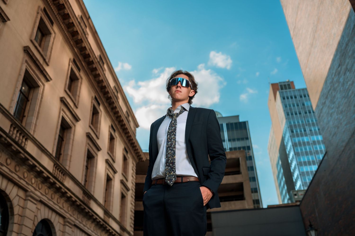 Stylish young man in suit and sunglasses posing between urban buildings under blue sky.