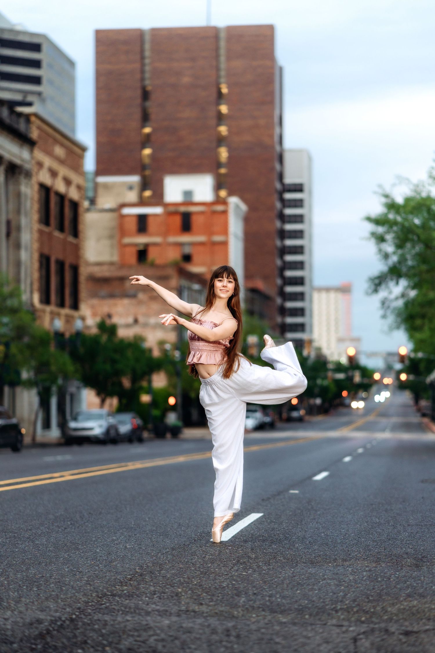 A ballet dancer performs an arabesque on pointe in white pants on an urban city street with tall buildings in the background.