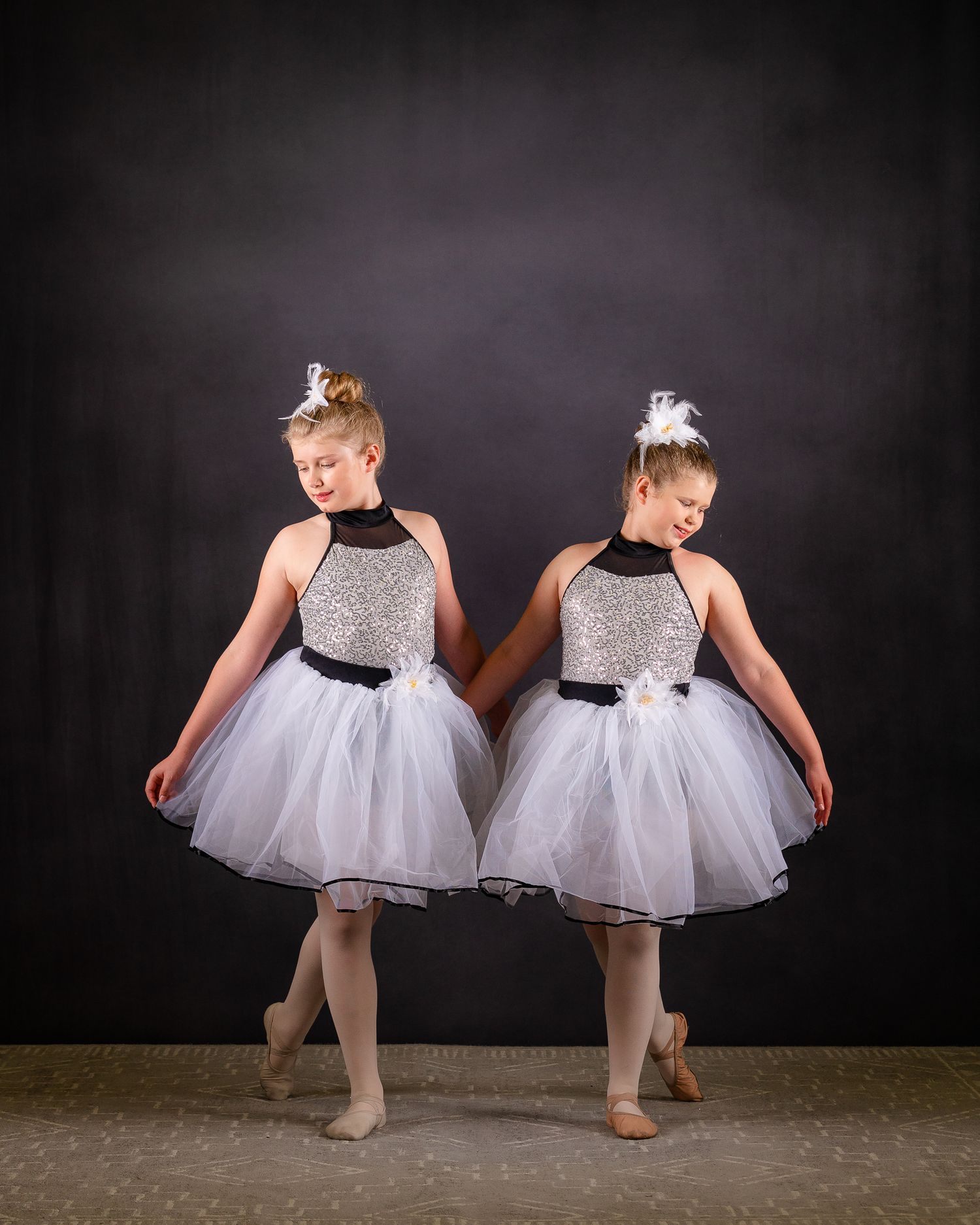 Two young ballet dancers in silver and white tutus posing together on dark stage.