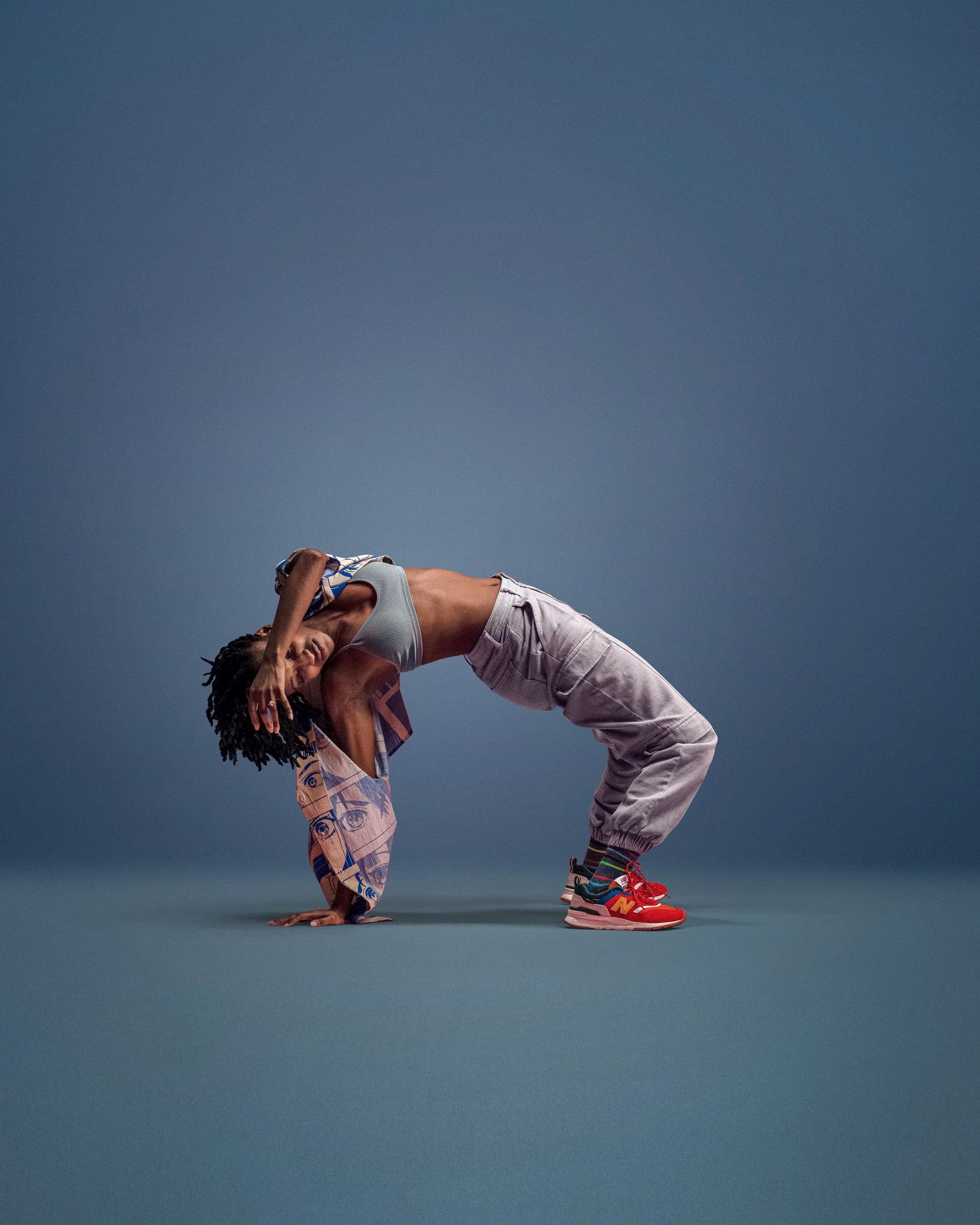 A dancer performs a dramatic backbend in a blue studio, wearing gray pants and red sneakers.