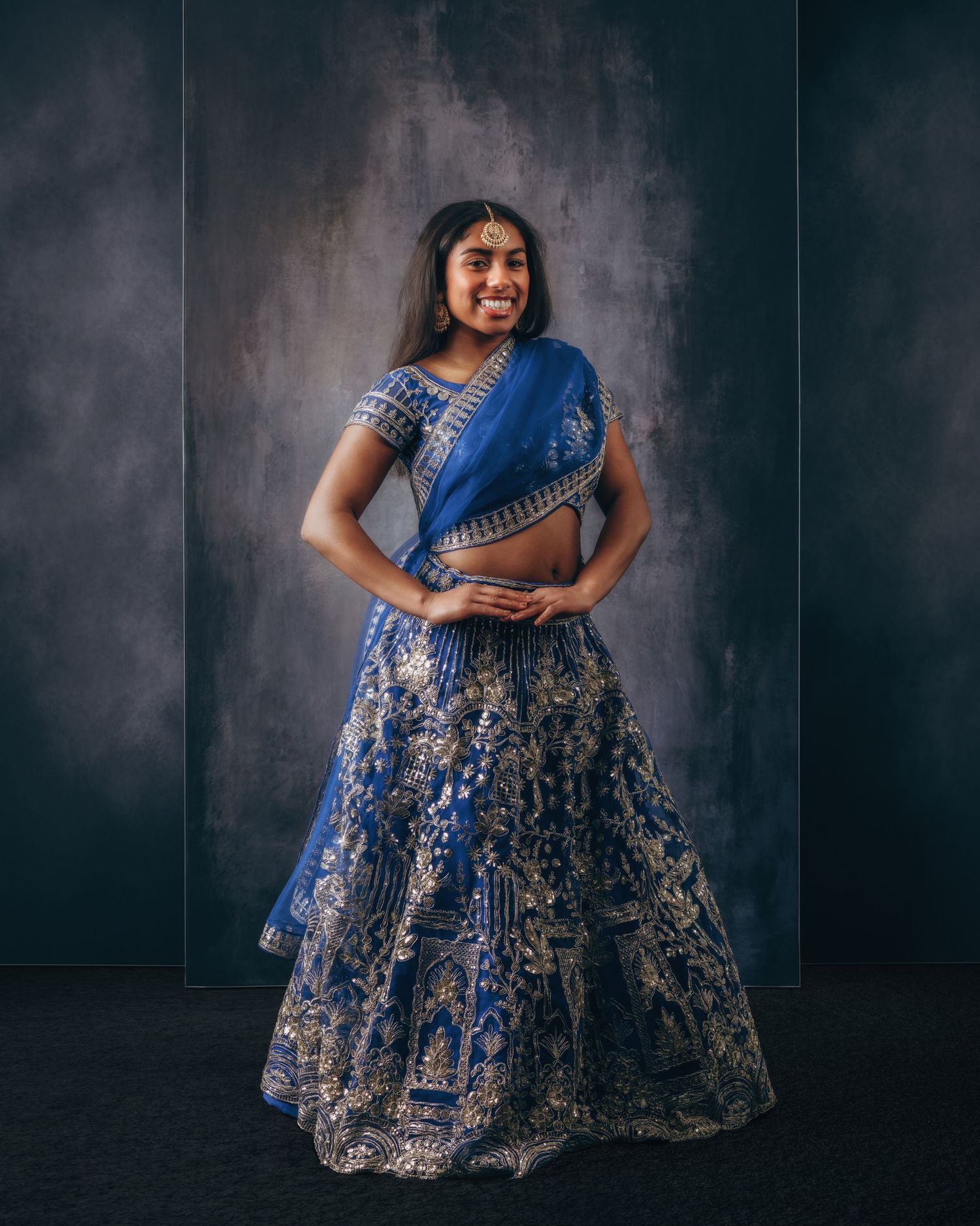 Young woman smiling in ornate royal blue and gold embroidered lehenga choli with dupatta for Indian occasion.