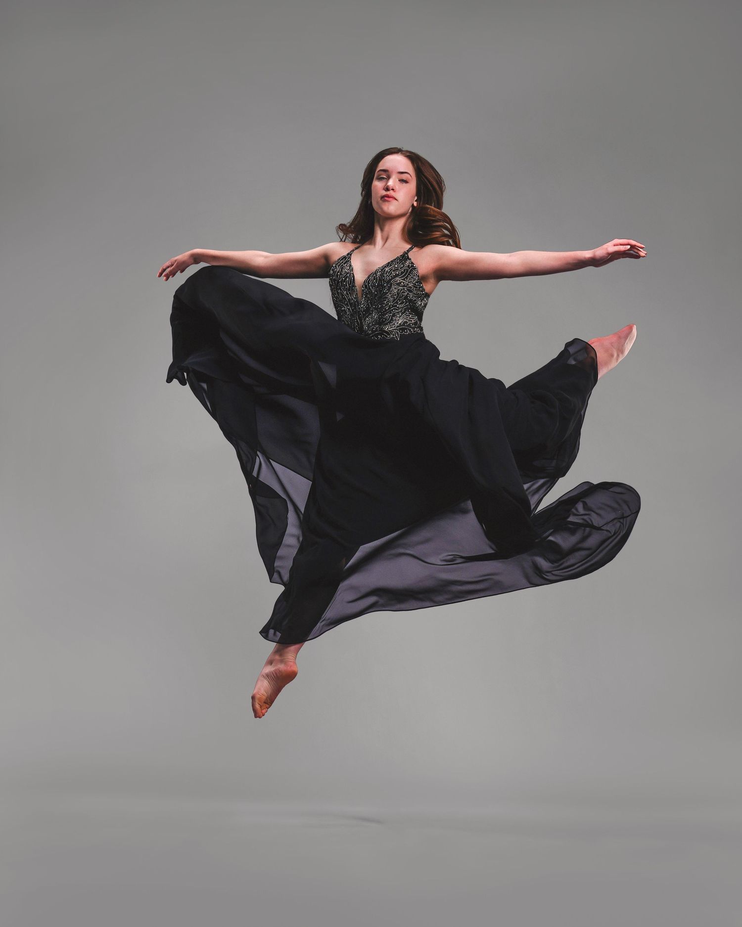 A dancer leaps gracefully in a flowing black dress, arms outstretched, against a gray studio background.