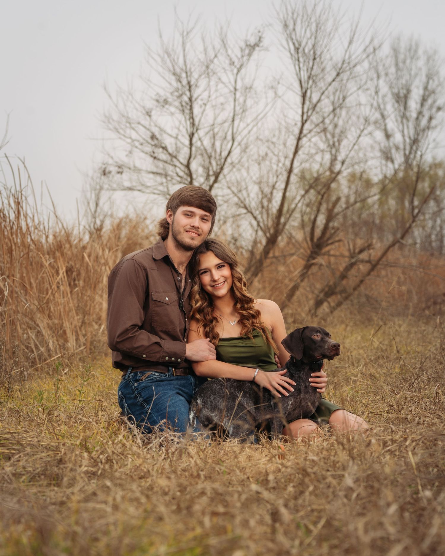Happy couple sitting together with their black dog in a dry autumn meadow.