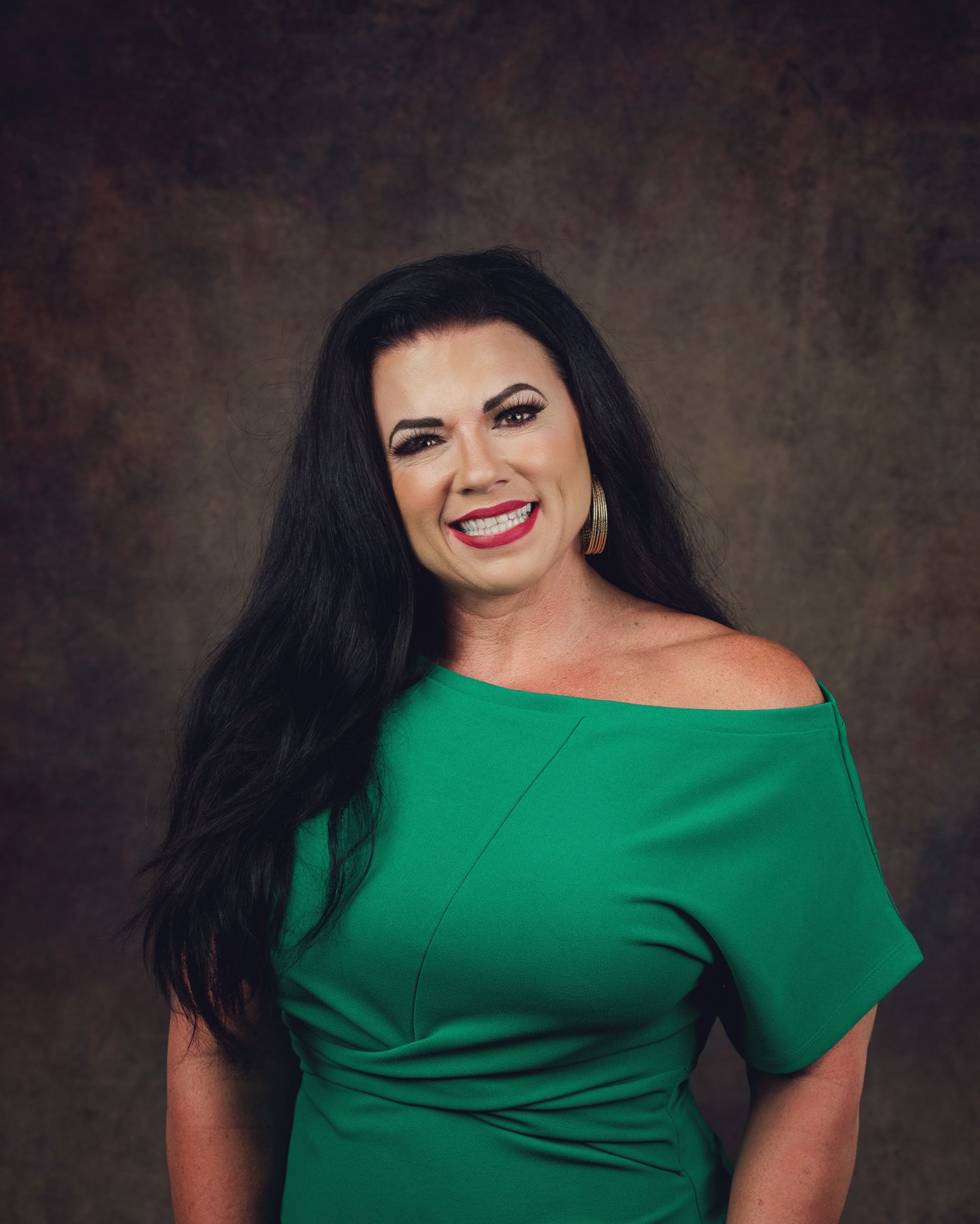 Smiling woman in green off-shoulder dress posing for professional portrait against brown backdrop.