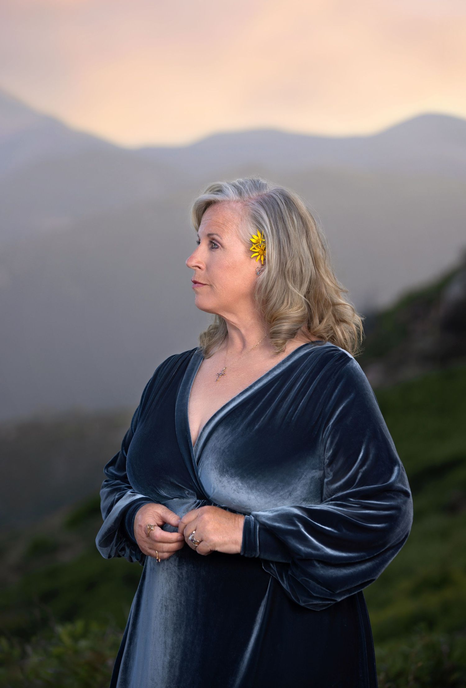 Elegant woman in blue velvet dress with yellow flower hair clip against mountain sunset.