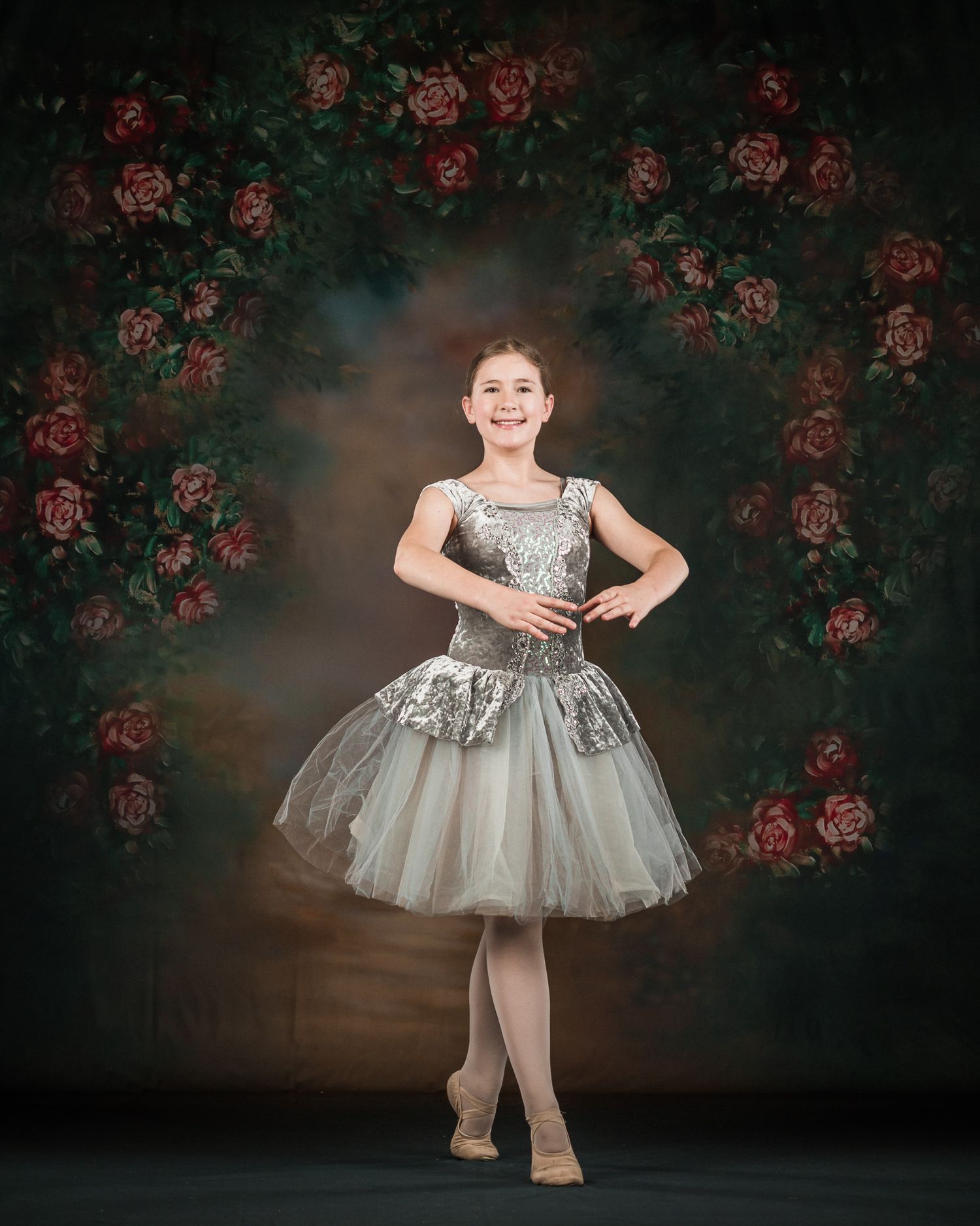 Young ballet dancer in silver tutu smiling before floral rose arch painted backdrop.
