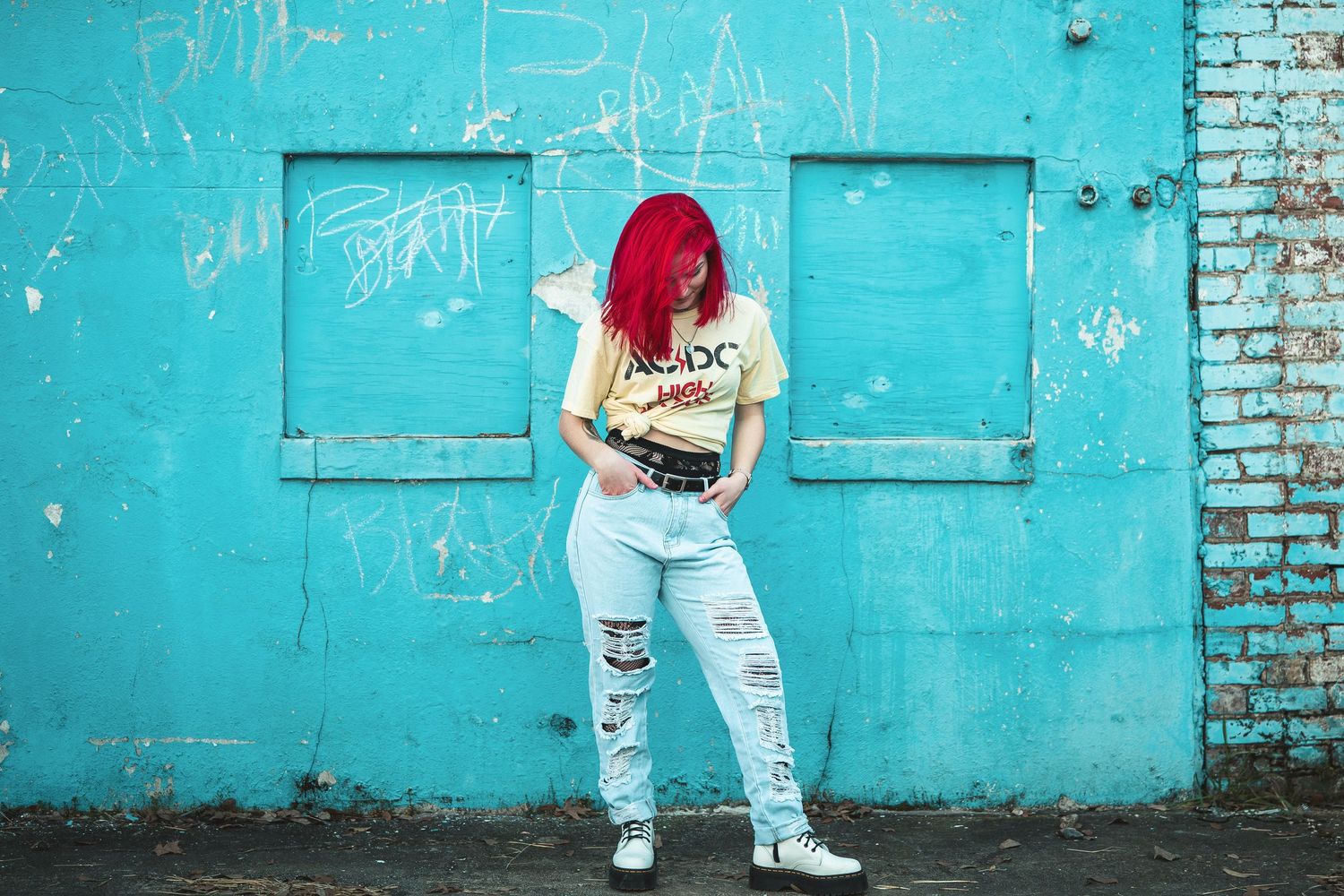 Red-haired woman in ripped jeans and yellow tee reads a book against a turquoise brick wall.