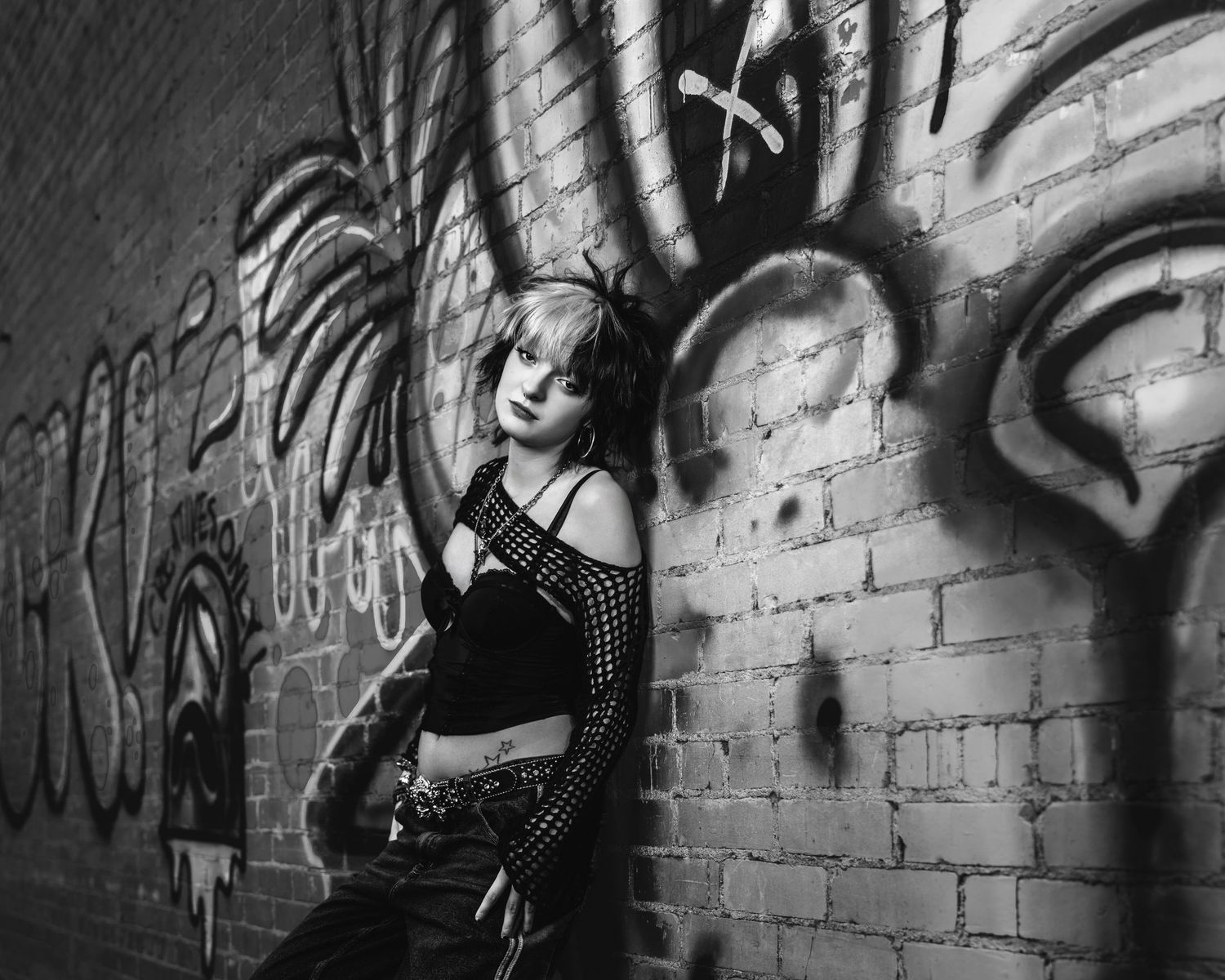 Black and white photo of a woman leaning against a graffiti-covered brick wall.