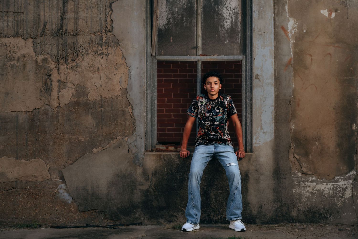 Young man in floral shirt and jeans poses confidently against a weathered urban wall with brick window.