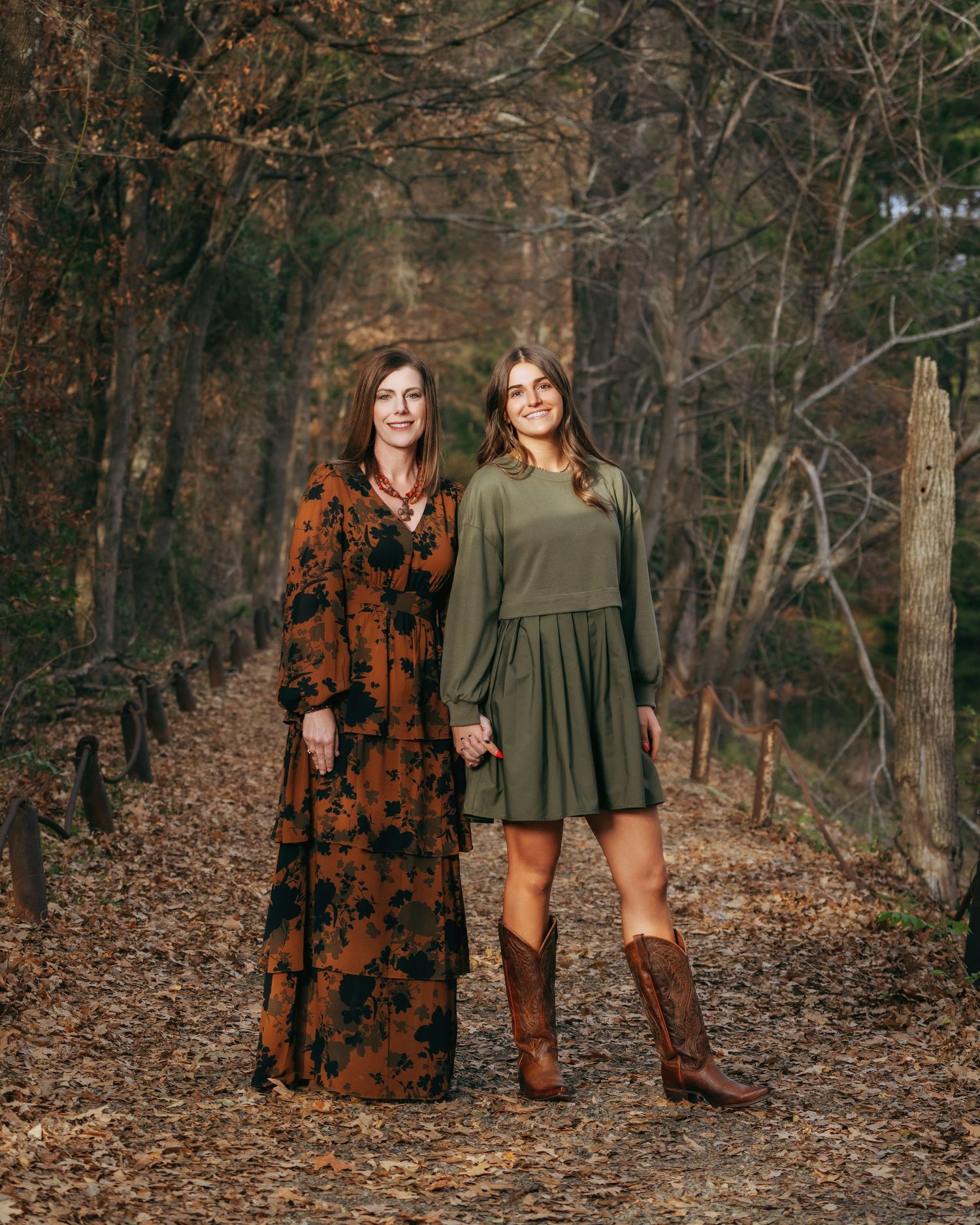 Two women holding hands on a leaf-covered forest trail, wearing green and floral dresses with cowboy boots.