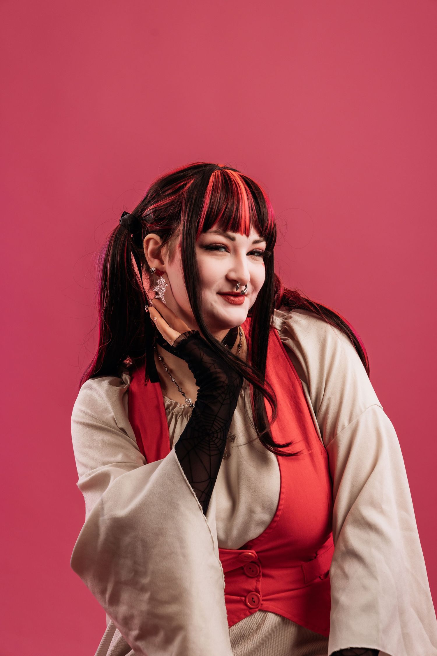 Woman in red top and white jacket with black gloves poses against a pink background with dark hair and red highlights.
