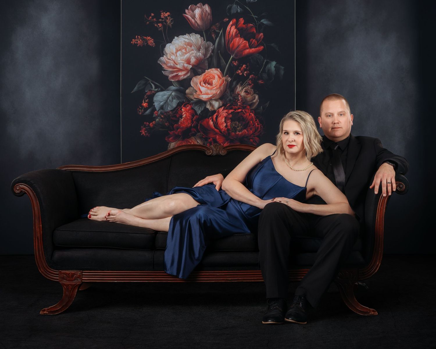 Elegant couple posing on black chaise lounge before floral backdrop, woman in blue gown, man in black suit.
