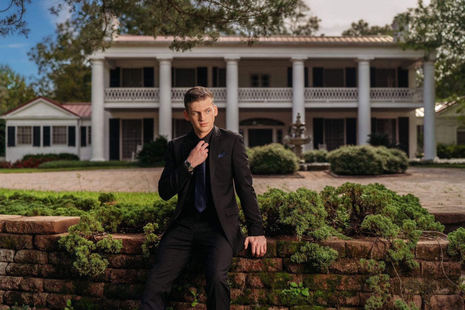 A well-dressed man in a dark suit adjusts his tie while leaning against a brick wall outside a white colonial mansion.