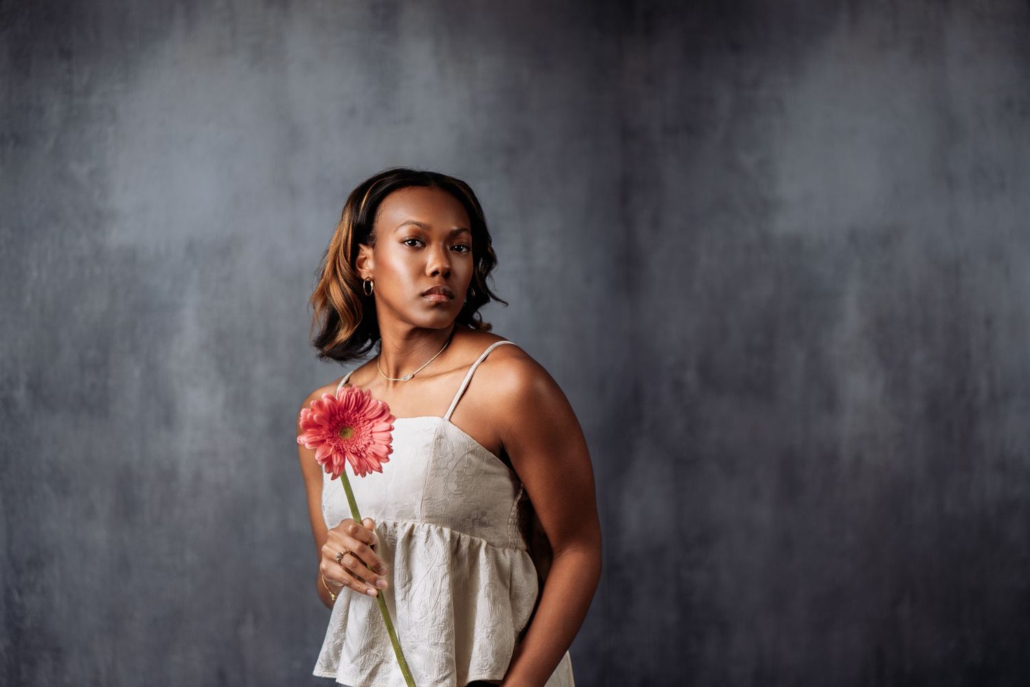 Young Black woman in a linen dress holds a pink gerbera daisy against a gray studio background.