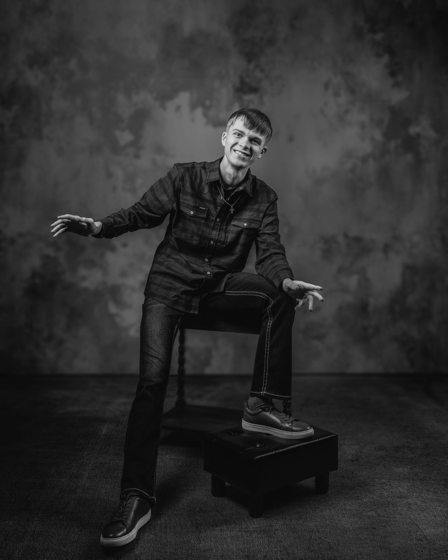 Black and white studio portrait of a man in dark denim sitting casually on a stool with a textured backdrop.