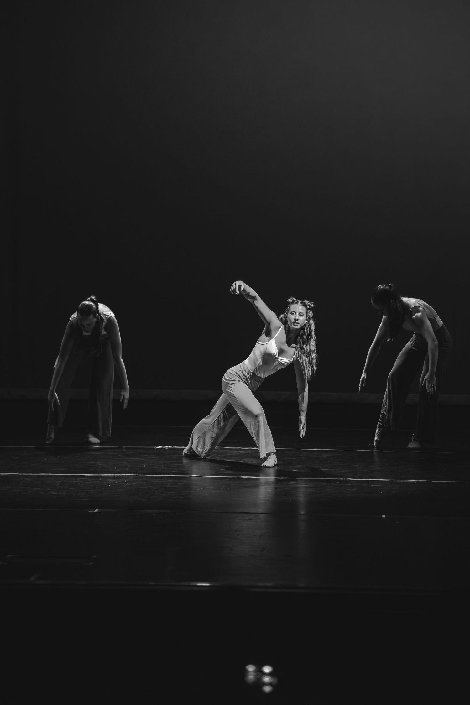 Black and white photo of three dancers performing on a dark stage, with one dancer in a dramatic lean pose.