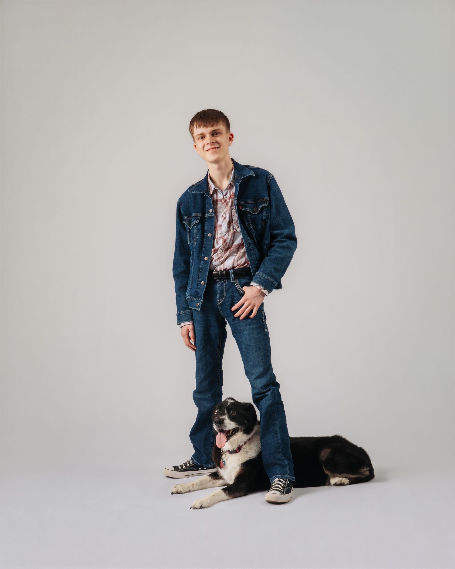 Young man in denim jacket and jeans stands beside a black and white Border Collie dog on gray background.