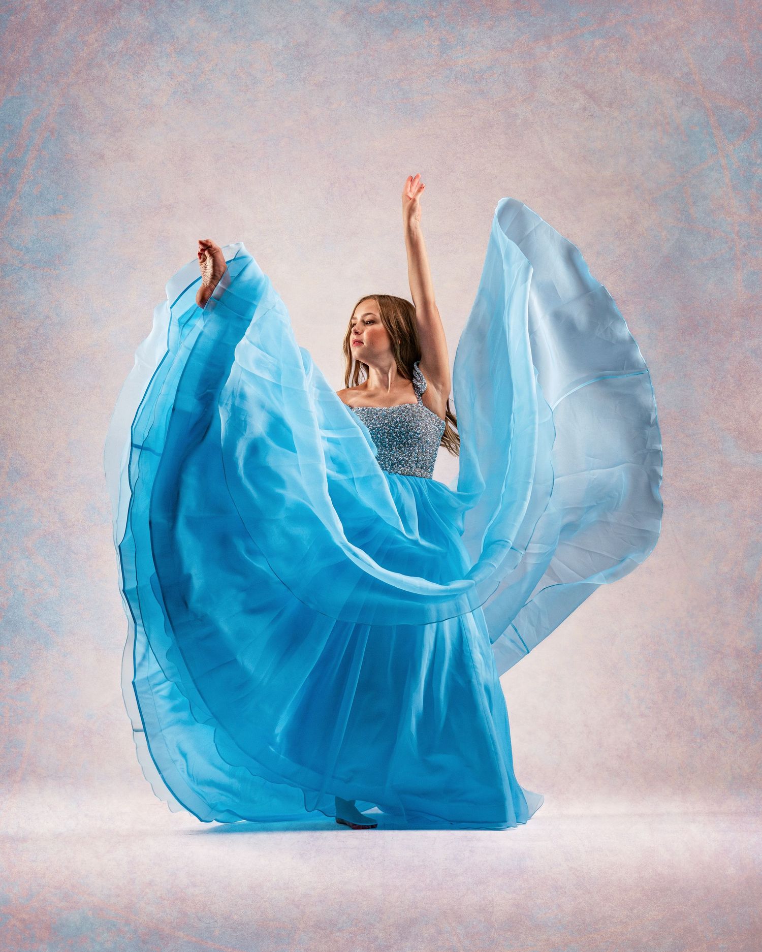 A dancer in a flowing turquoise gown twirls gracefully with arms raised against a soft pink background.