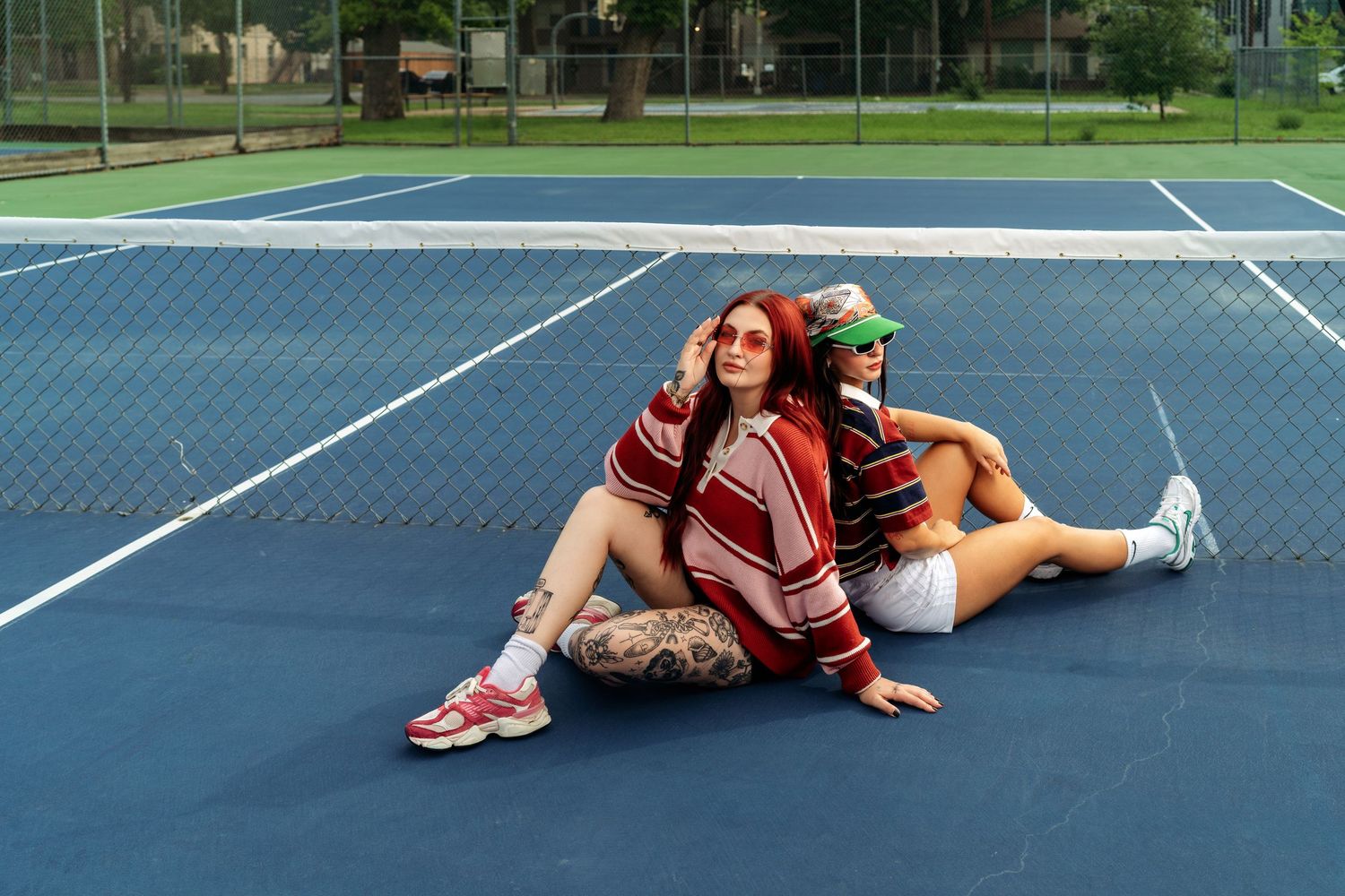 Two women sit together on a blue tennis court, one wearing a red sweater and the other in white shorts.