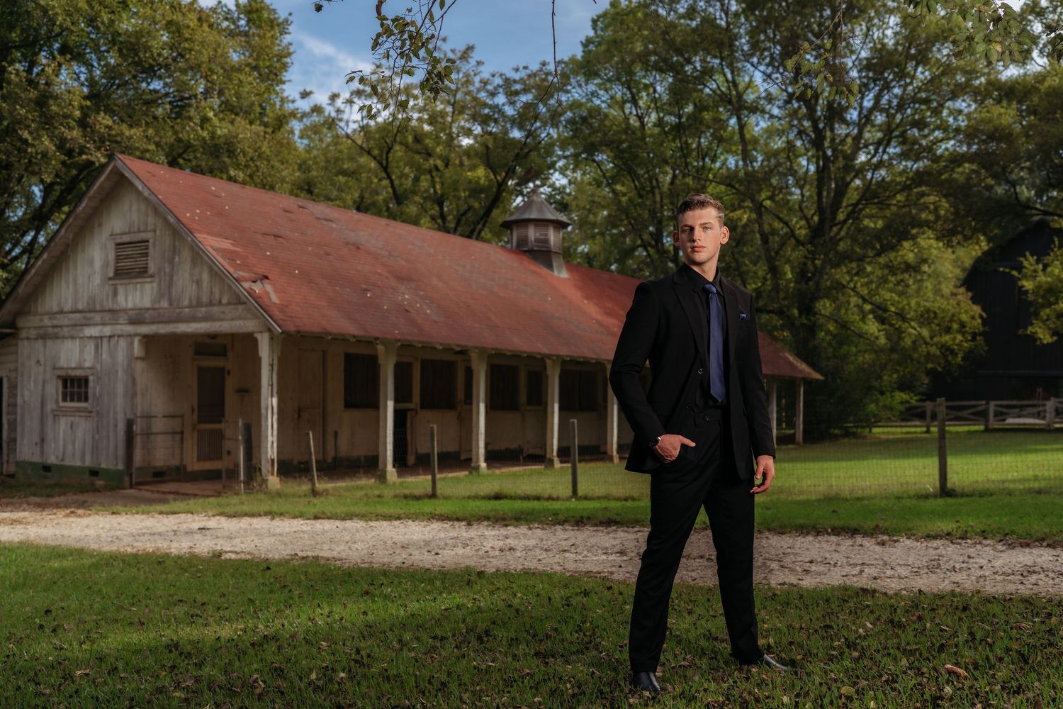 A man in a black blazer stands on a grassy field in front of a rustic wooden barn.