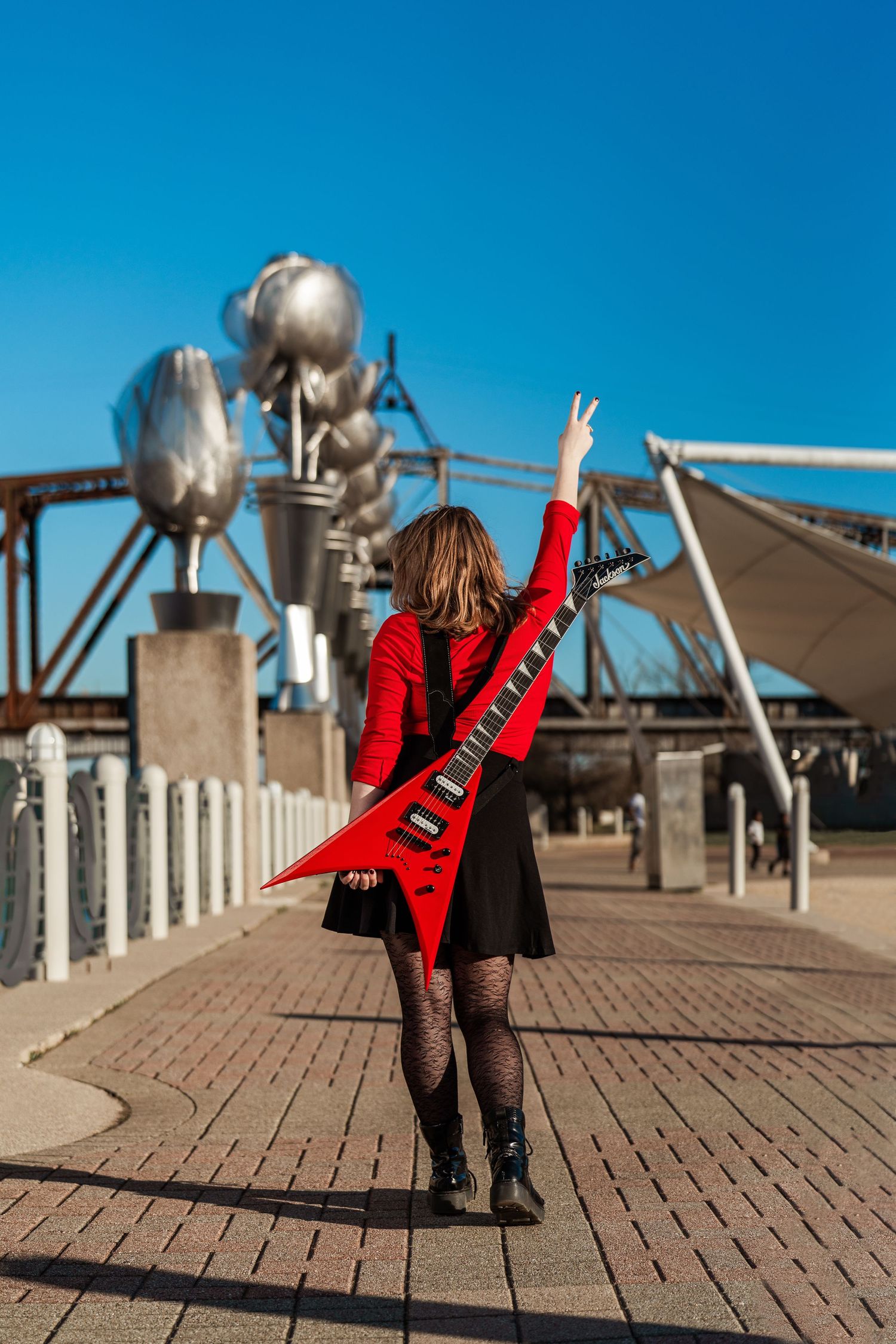 Woman in red jacket carrying a red electric guitar on a bridge, making a peace sign on a sunny day.