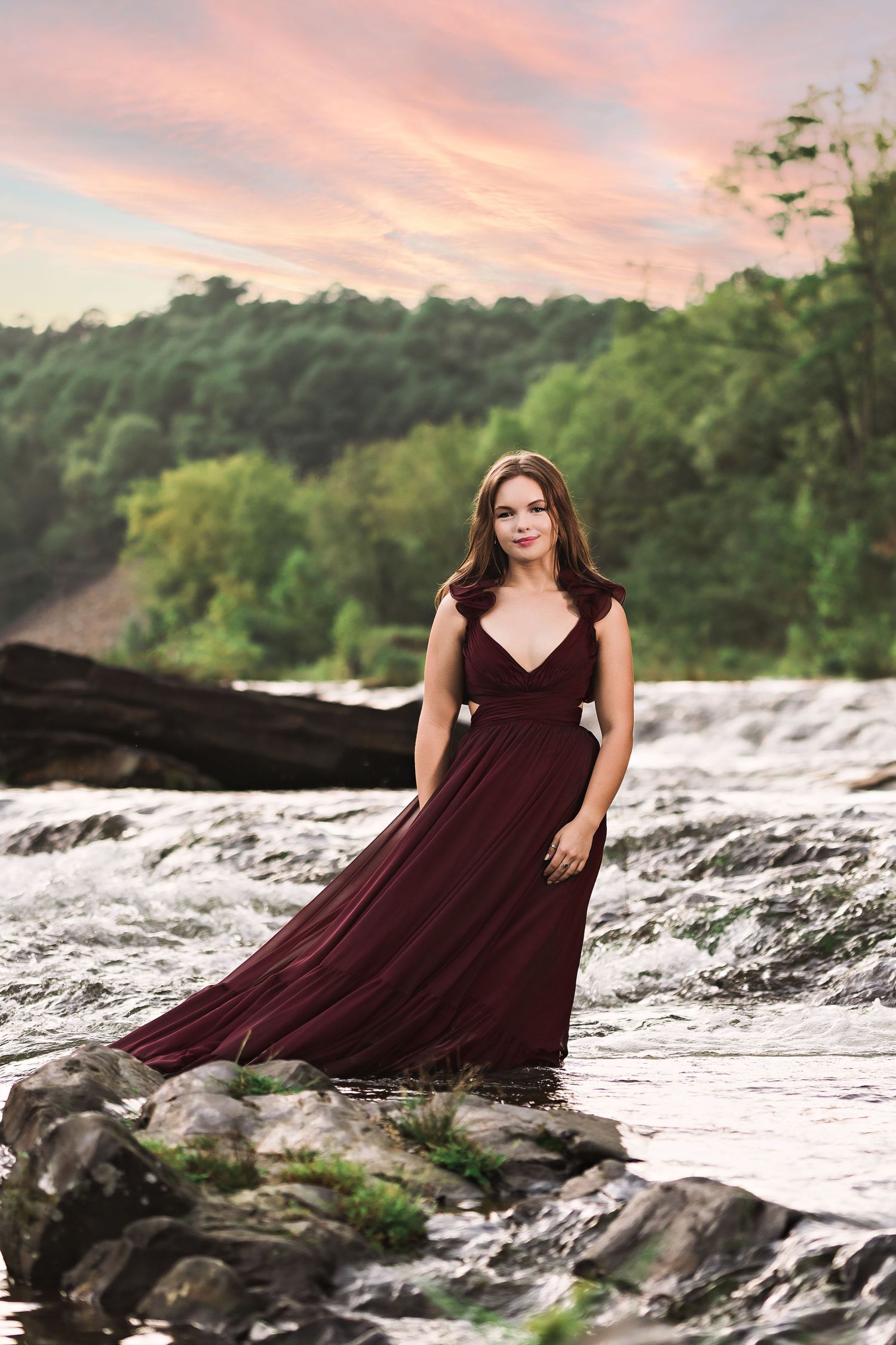 Woman in flowing burgundy gown stands on rocks by a rushing river at sunset with lush green trees behind her.