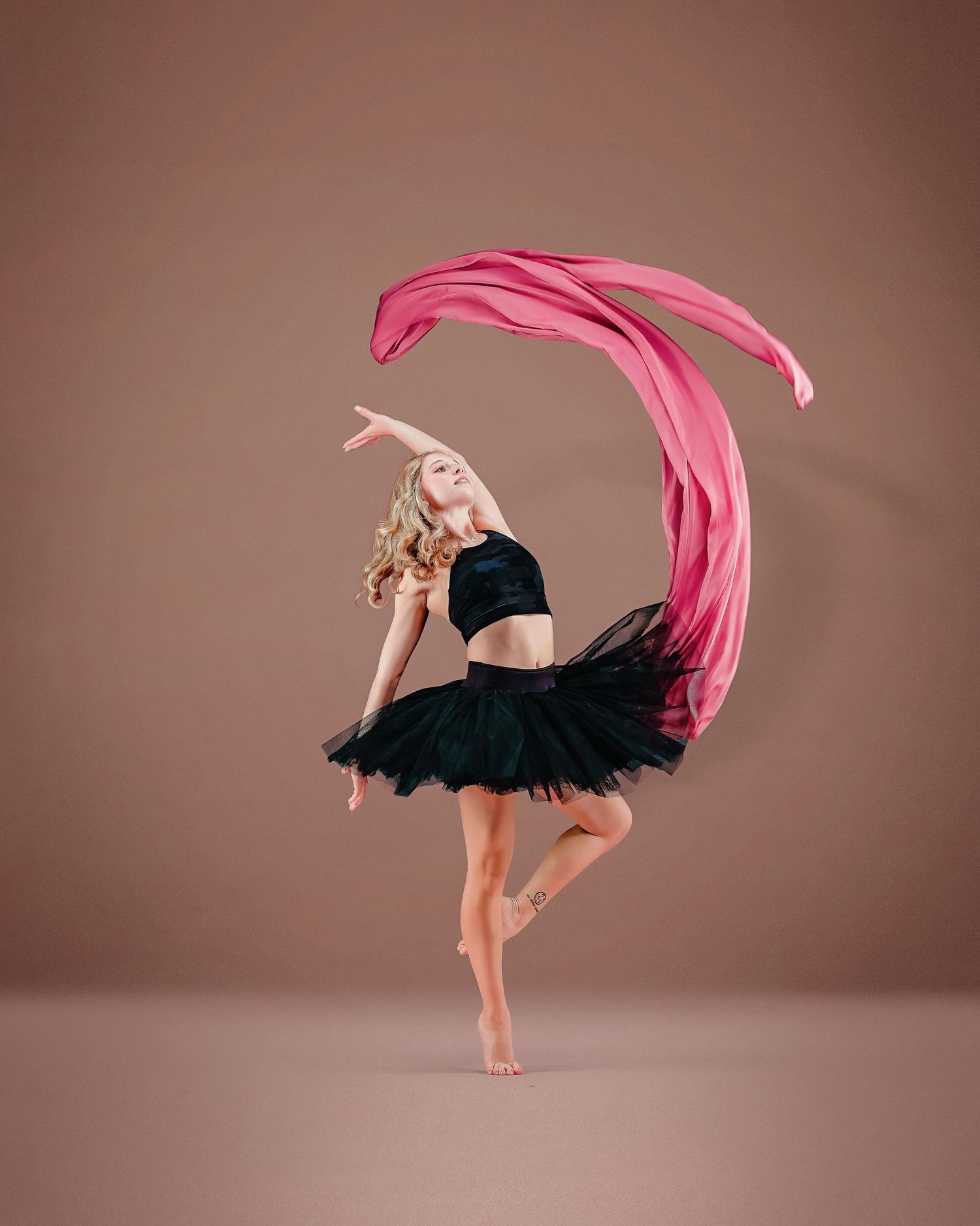 Blonde dancer in black tutu leaps gracefully with flowing pink fabric swirling overhead in a studio.
