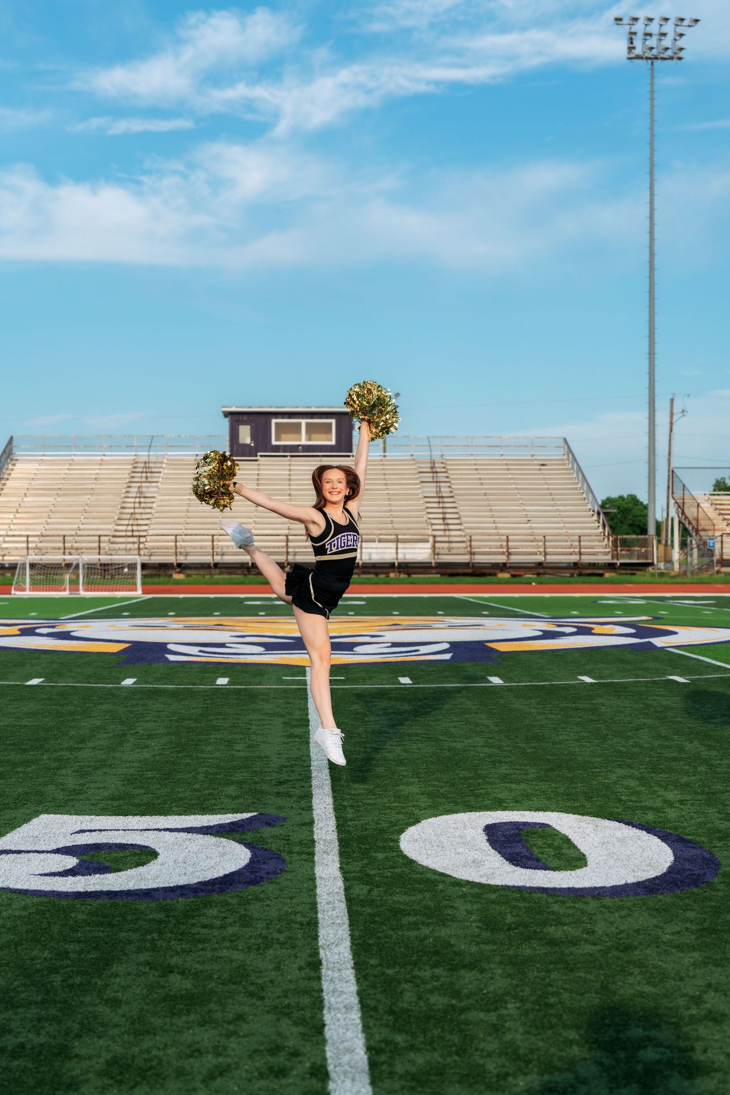 Cheerleader in black uniform poses with gold pom-poms on the 50-yard line of a football field under blue skies.